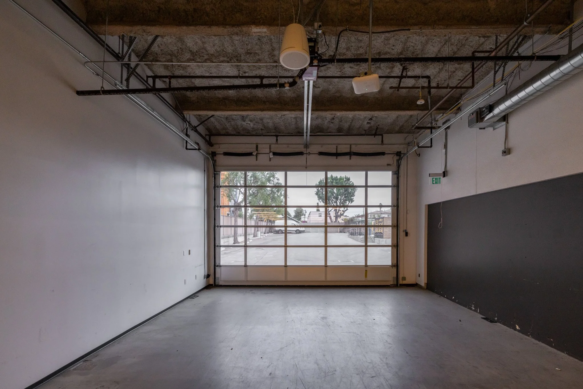 Empty industrial garage or workshop with large roll-up glass door, concrete floor, white and black walls, and exposed ceiling pipes and ducts