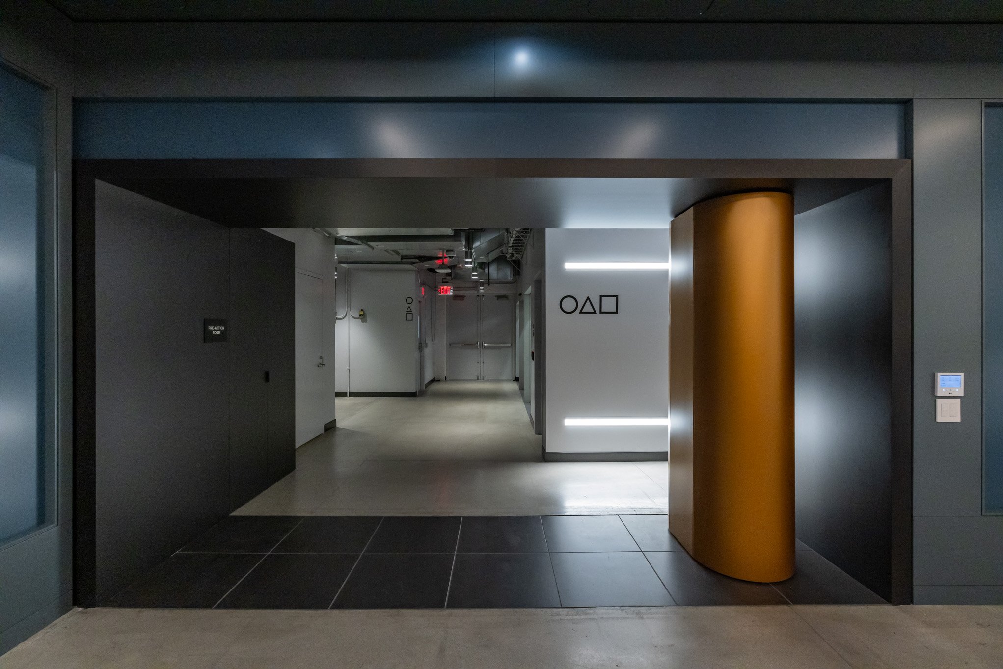 Modern office corridor with black walls, gray doors, and decorative symbols of a circle, triangle, and square on the walls, illuminated by subtle lighting and featuring a golden cylindrical column on the right.