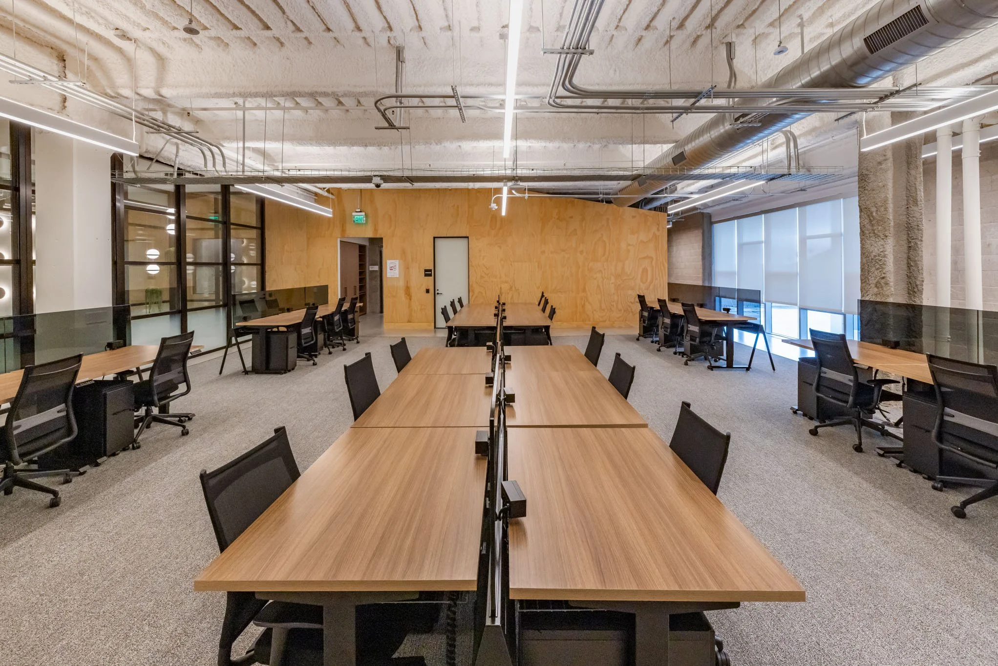Modern conference room with long wooden tables, black office chairs, and large windows with blinds, in an industrial-style interior.