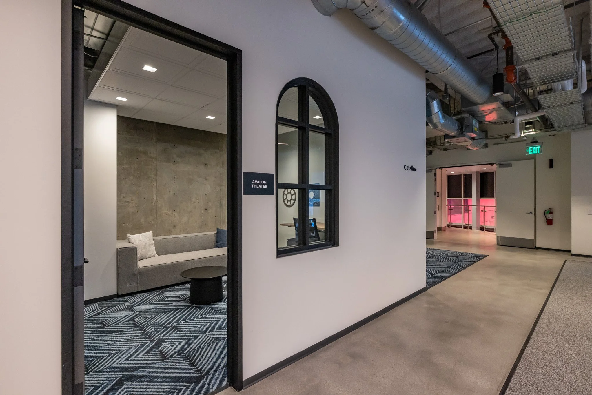 Interior view of a modern building hallway with an open door leading to a waiting area with a gray couch and a small black table. There is a black-framed window on the wall with the sign 'Avalon Theater,' and the walls are white. The ceiling has expo
