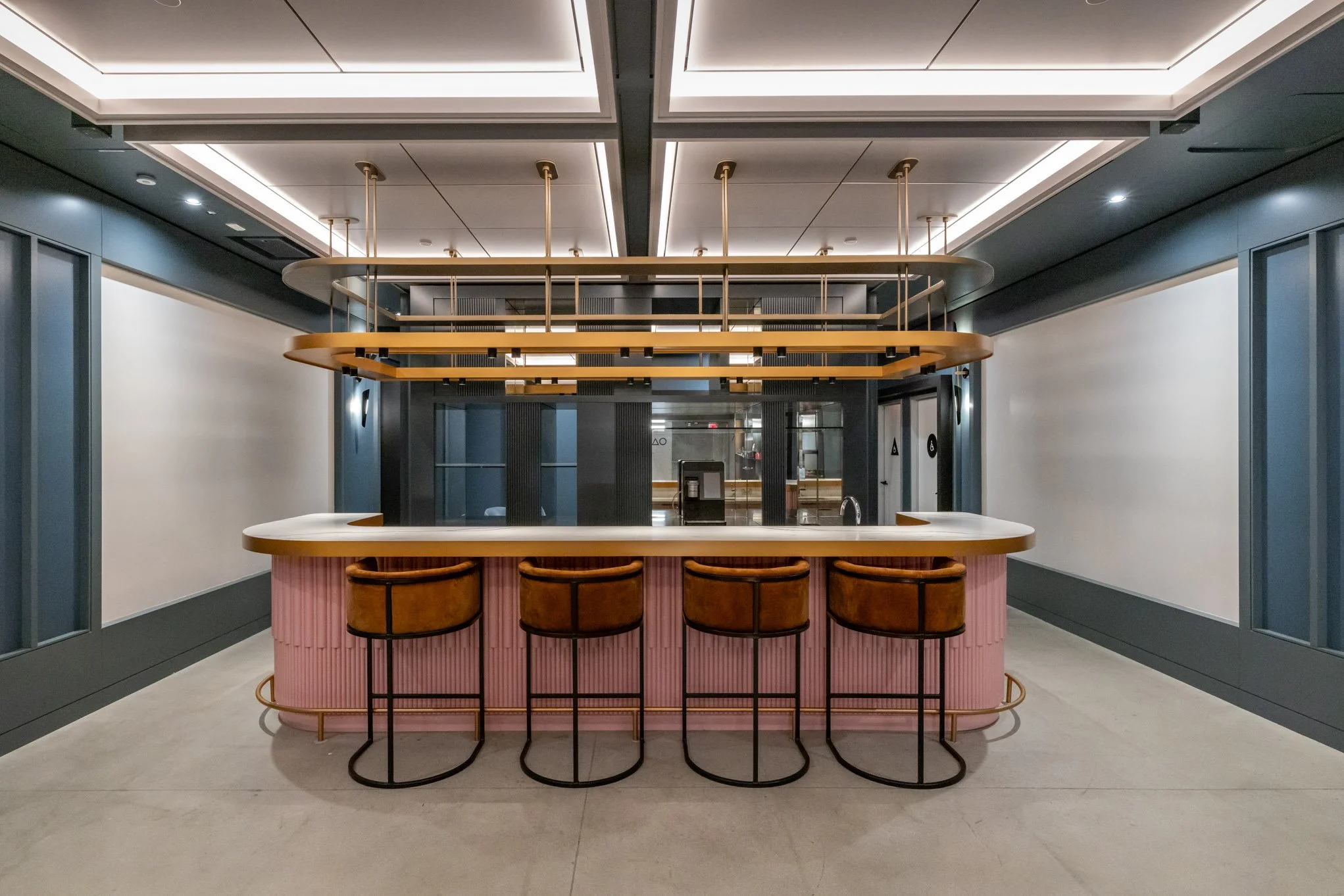 Modern hotel reception area with a curved pink desk, four leather bar stools, overhead lighting fixtures, and an elevator in the background.