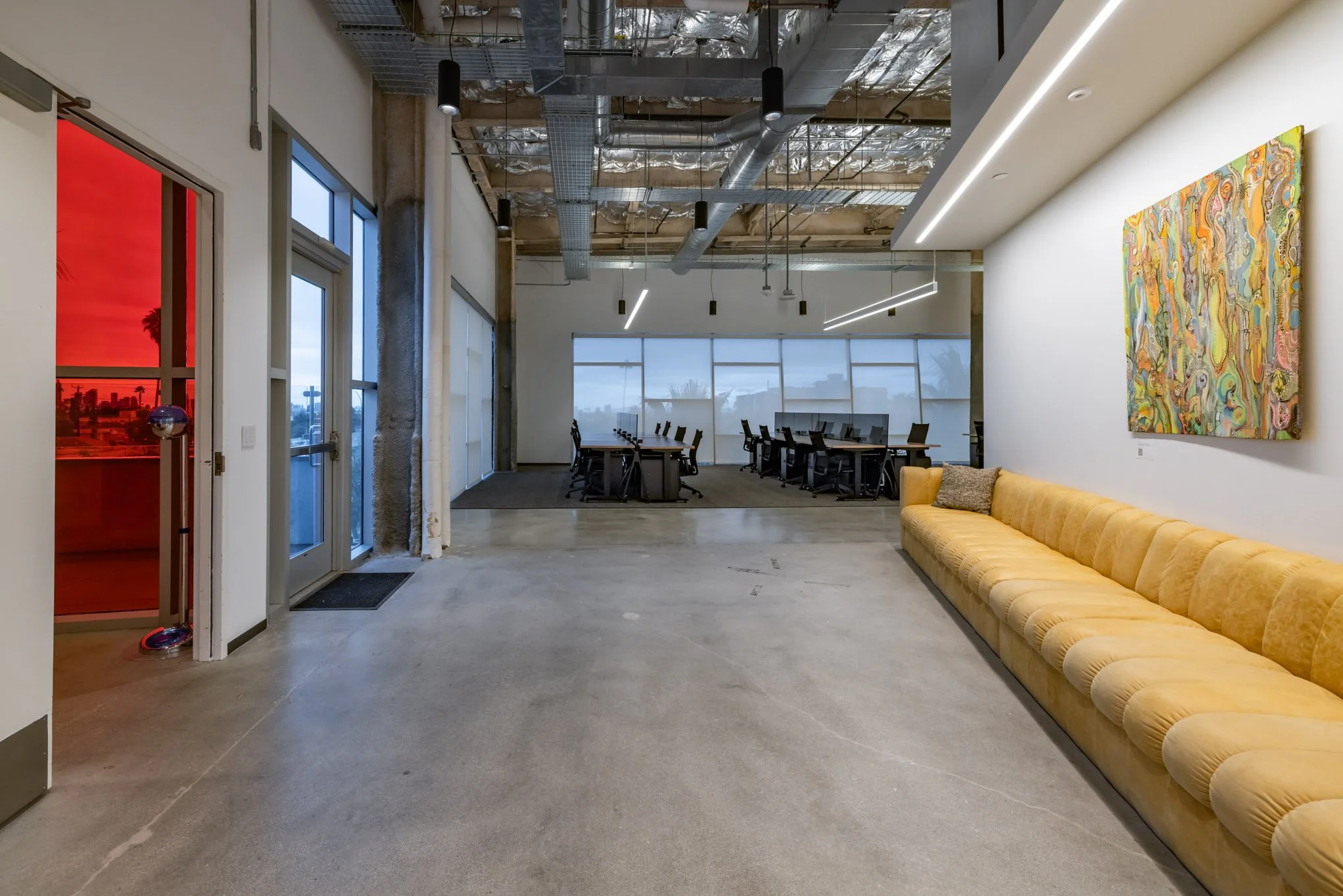 Modern office interior with a long yellow sofa, colorful abstract wall art, conference table and chairs, large windows, and exposed ceiling with ductwork and lighting fixtures.