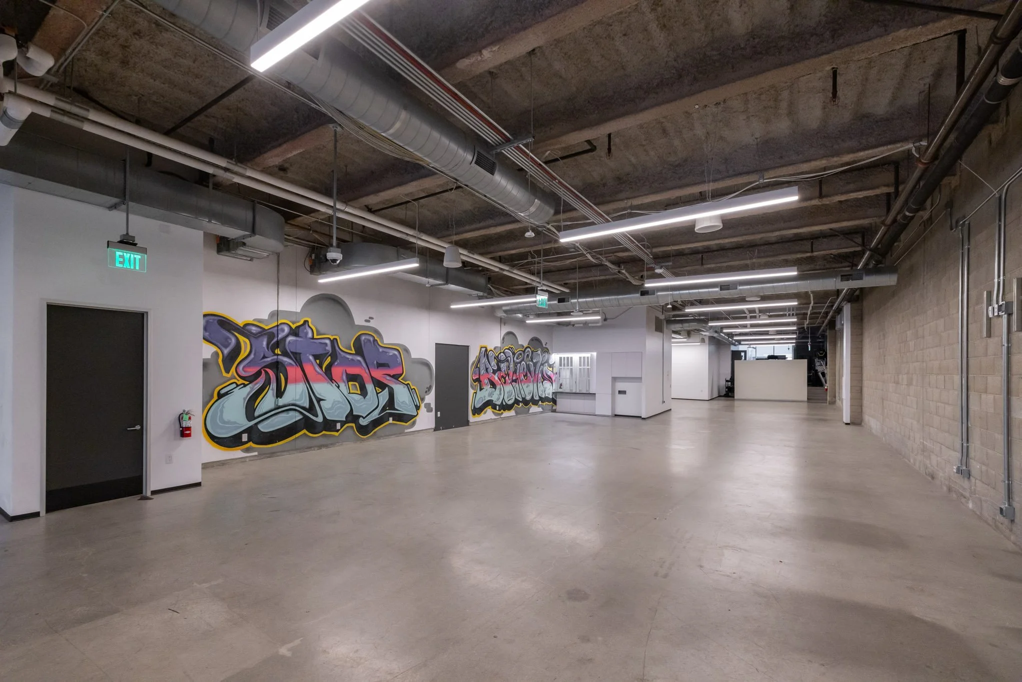 Empty industrial-style room with graffiti art on the white wall, exposed ductwork, and lighting fixtures.