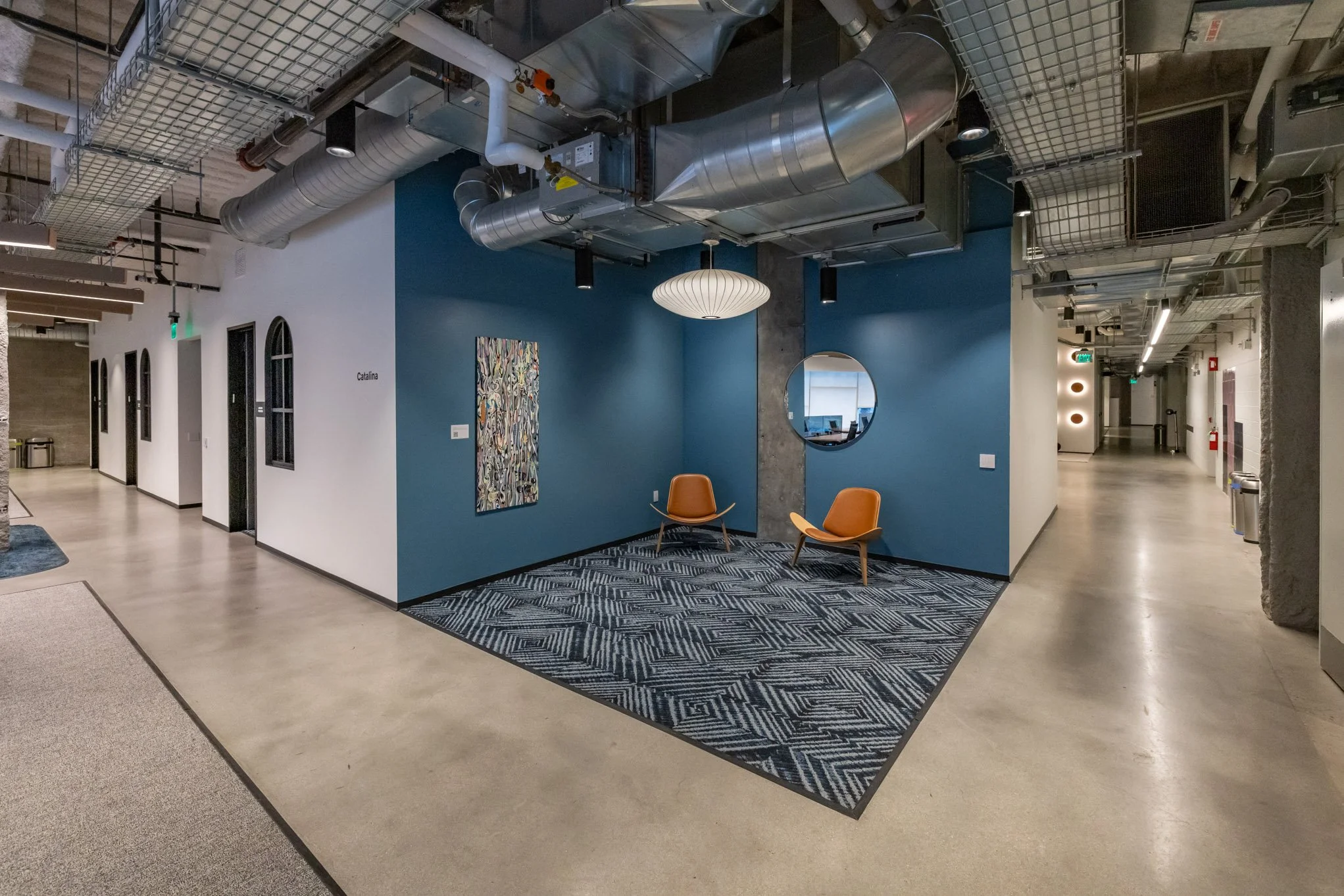 Modern office hallway with a small lounge area featuring two orange chairs, a blue accent wall with artwork and a round mirror, industrial ceiling with exposed ductwork, and additional hallway extending into the distance.