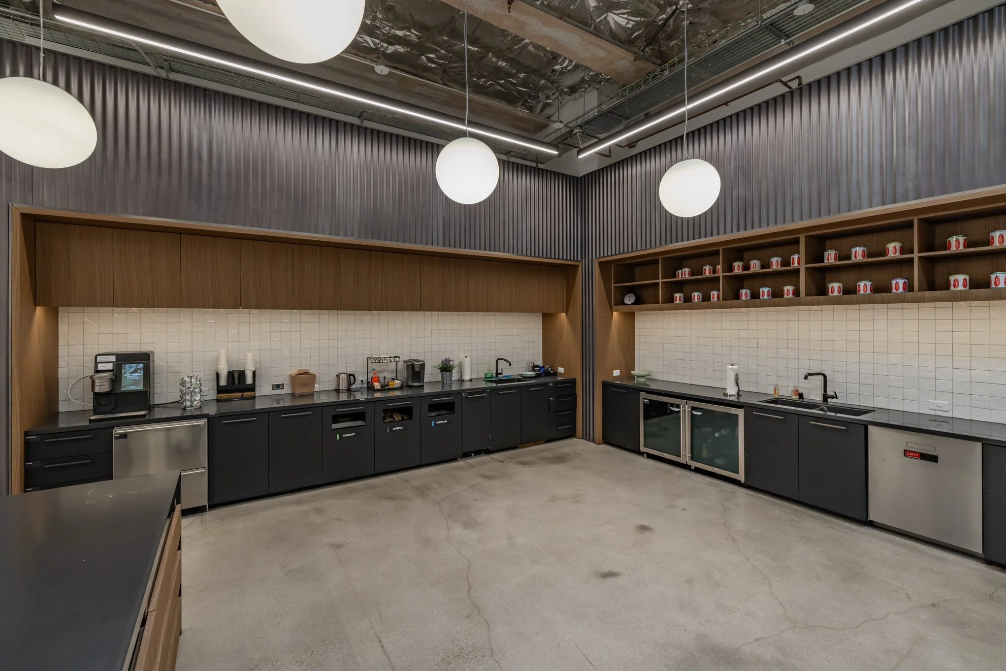 Modern coffee break area with black countertops, white tiled walls, and open wooden shelving. Contains a coffee machine, cups, and small appliances, with a concrete floor and hanging globe lights.