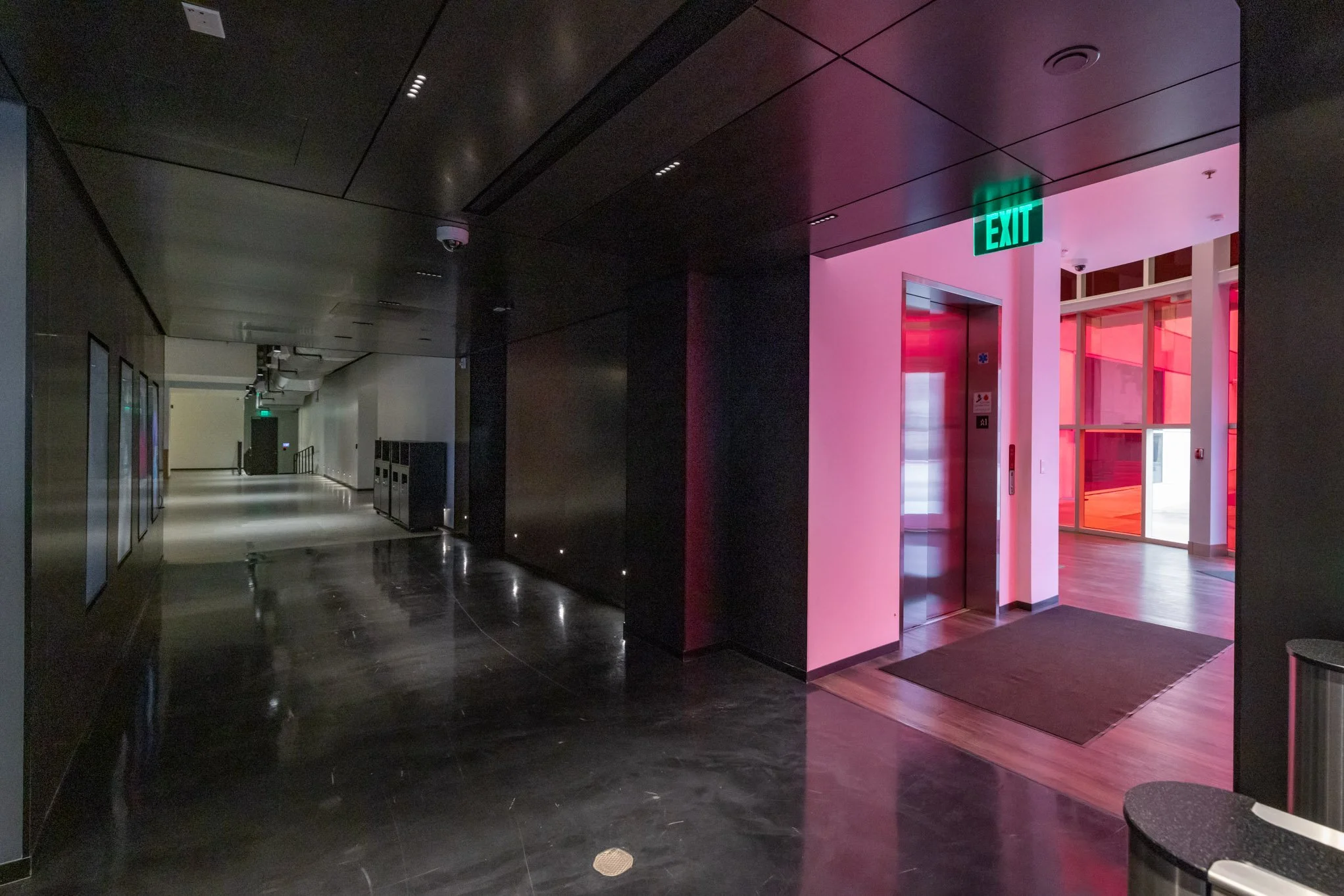 Interior hallway with dark flooring and walls, illuminated by small stage lights near the floor, leading to an elevator with a pink and red light reflection, and a green exit sign above the elevator door.