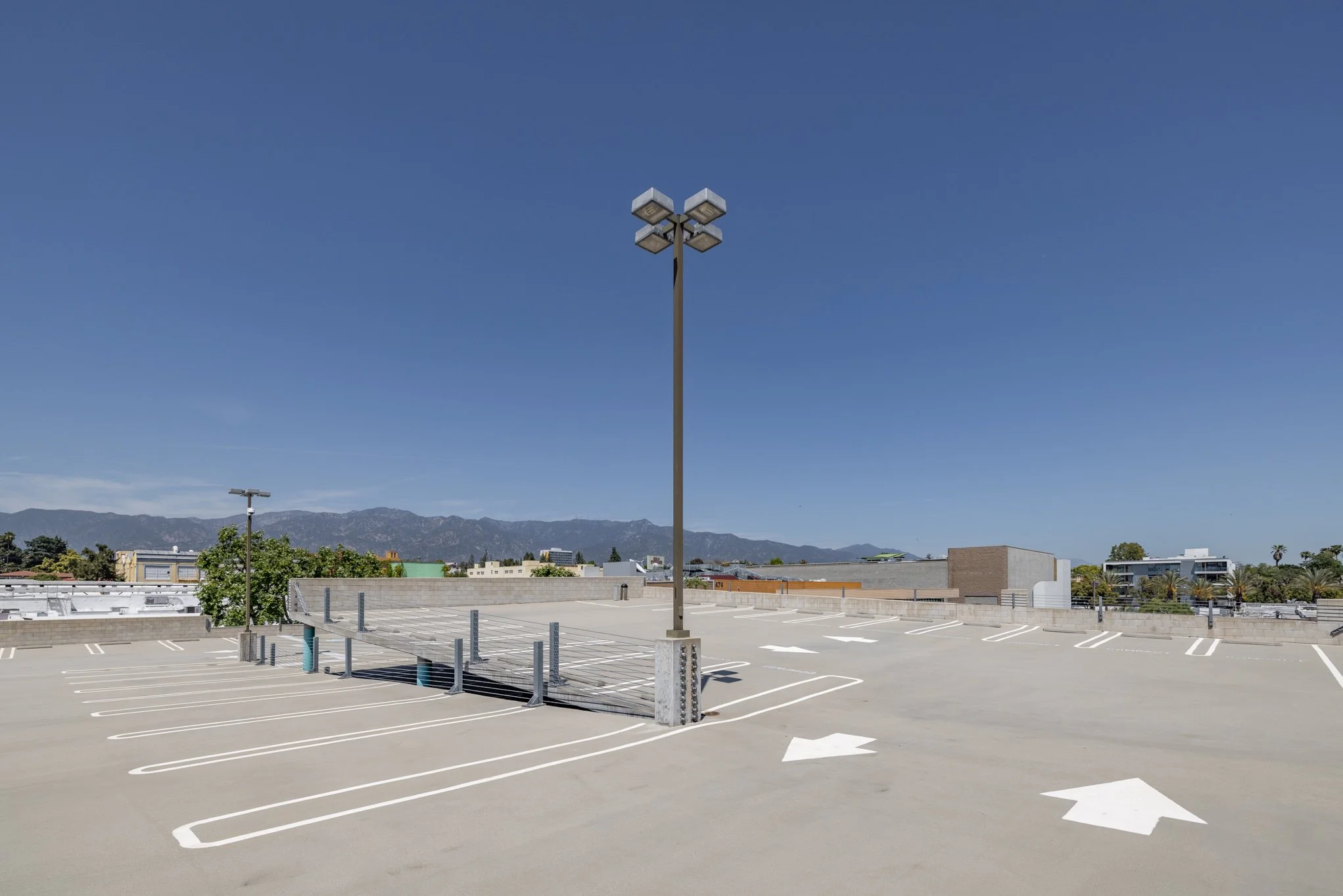 Empty rooftop parking lot under a clear blue sky with mountain range in the background, featuring a tall street light and white painted parking lines and arrows.