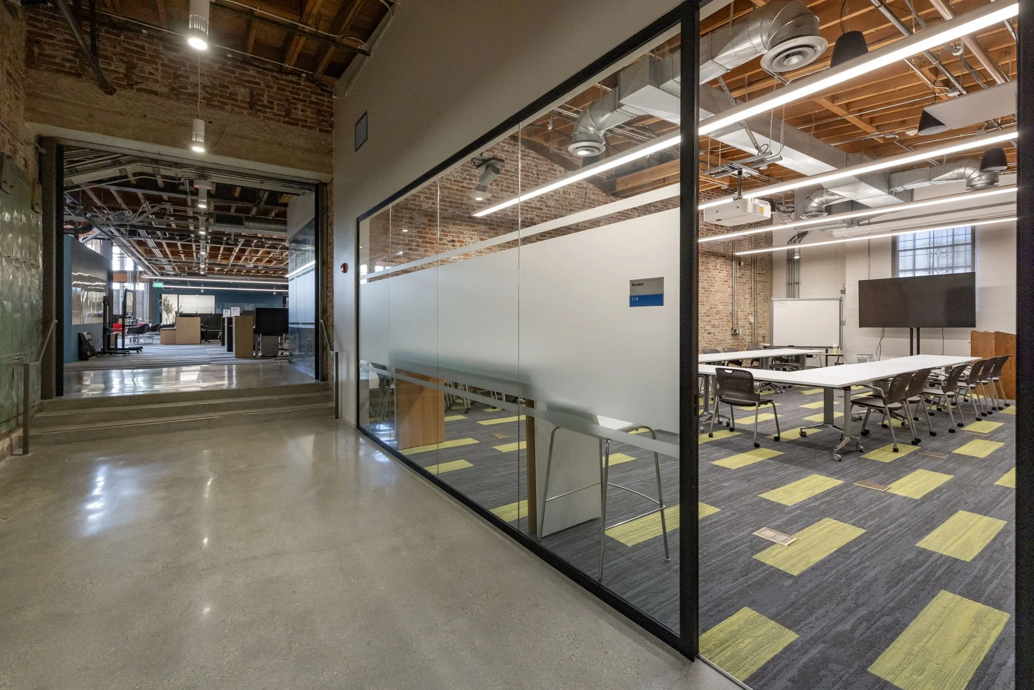 Modern office conference room with glass wall, white tables, black chairs, brick walls, and ceiling with exposed ductwork.