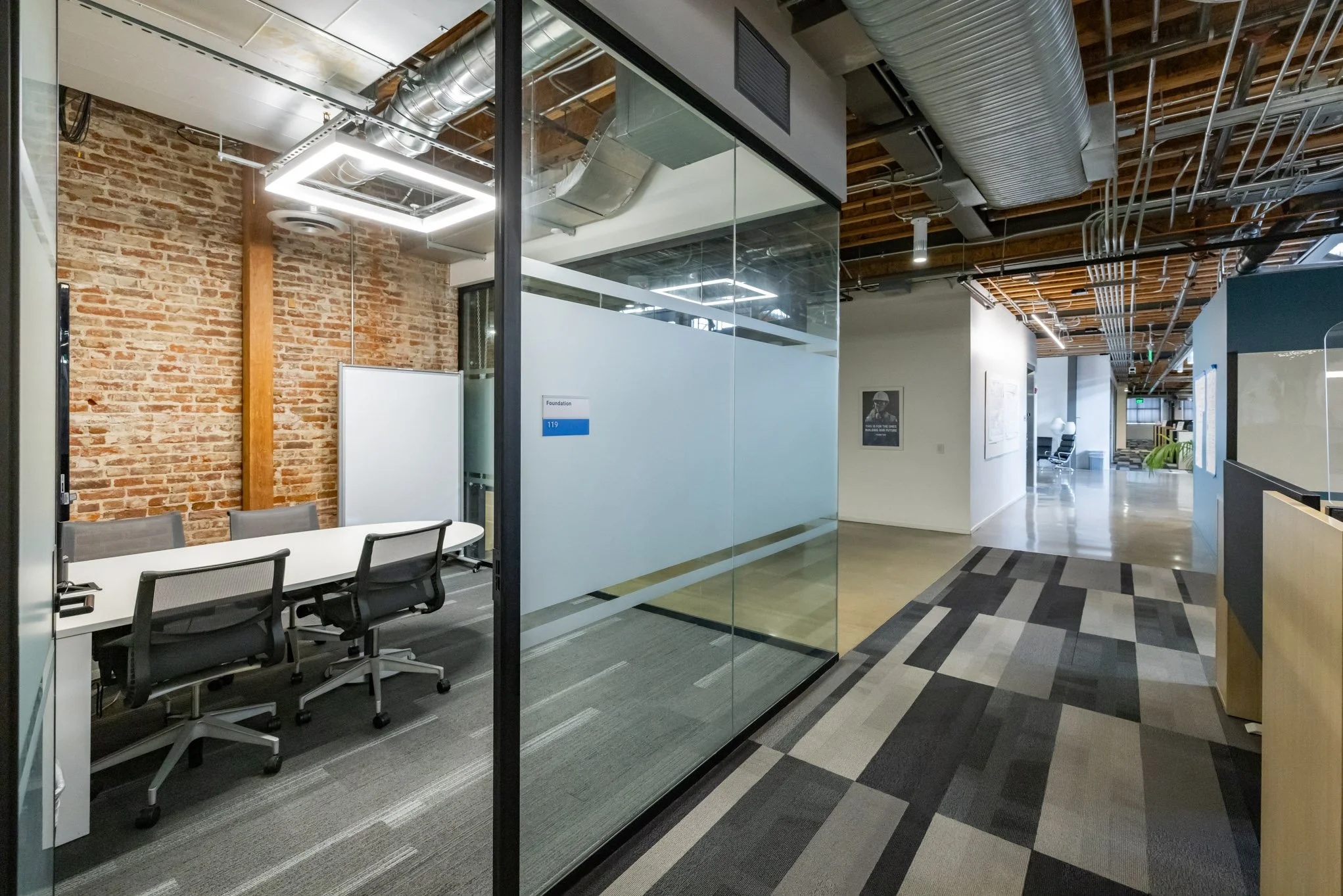 Modern office hallway with glass-walled meeting room on the left, featuring a brick wall, a wooden support beam, black office chairs around a white table, and silver ductwork and LED lighting on the ceiling. The hallway has a colorful, geometric patt