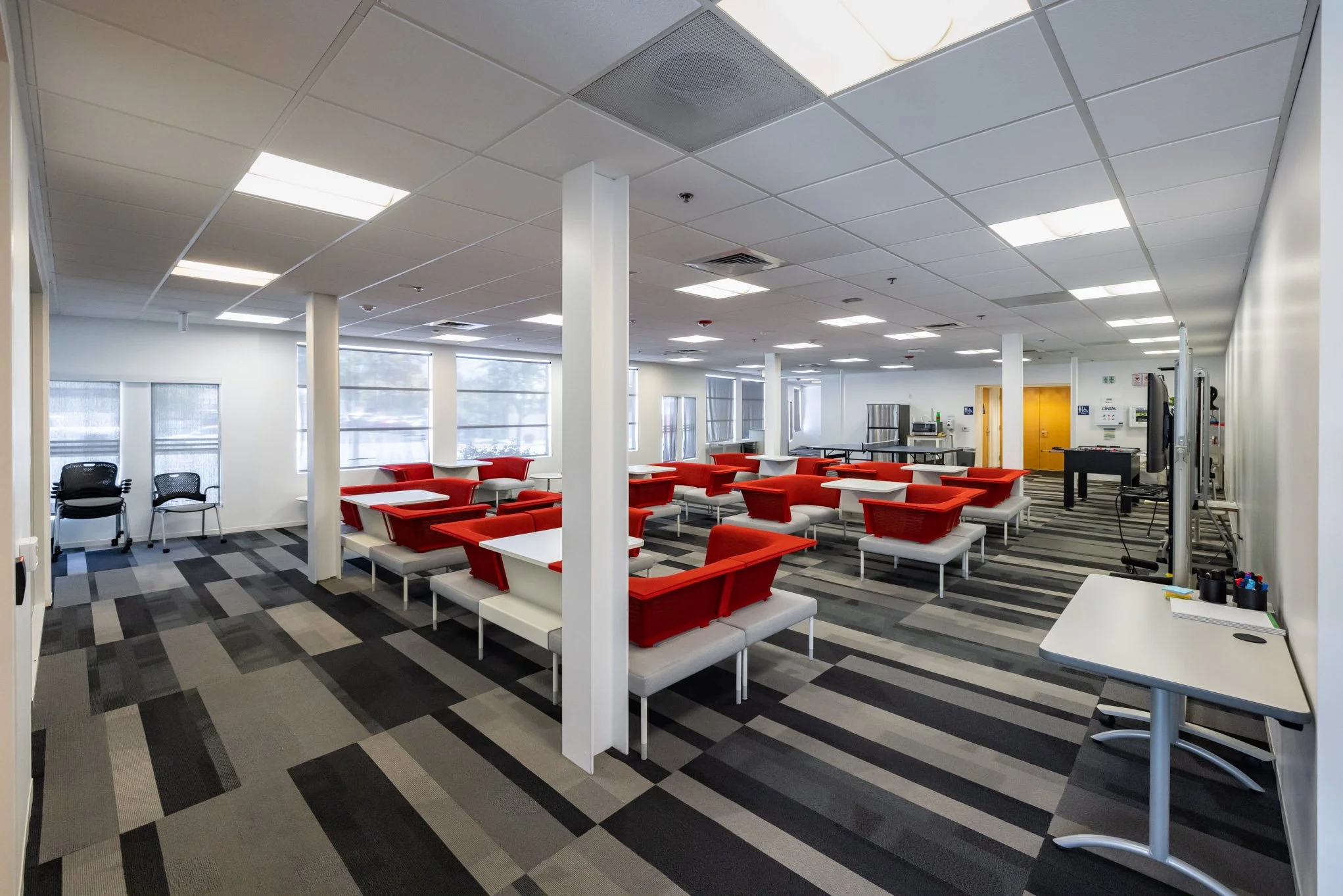 Empty cafeteria with white tables, red and white seating, and large windows, featuring a patterned gray carpet, white walls, and ceiling lights.