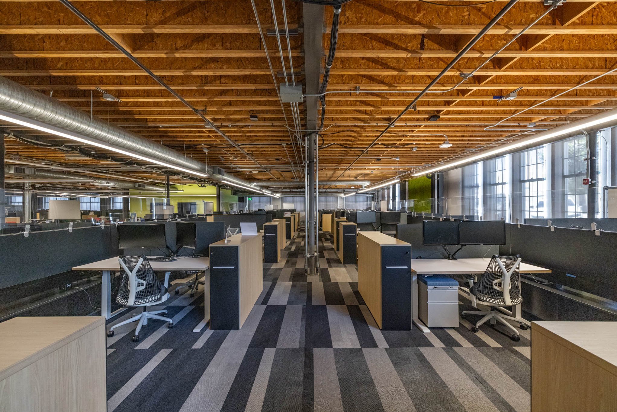 Empty modern office with rows of desks, black monitors, white office chairs, large windows, and a wooden ceiling.