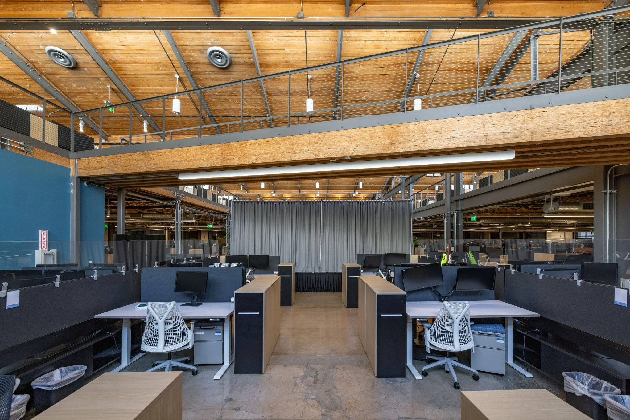 Open office workspace with multiple desks and monitors, a stage with a curtain in the background, and modern wooden and metal interior design.