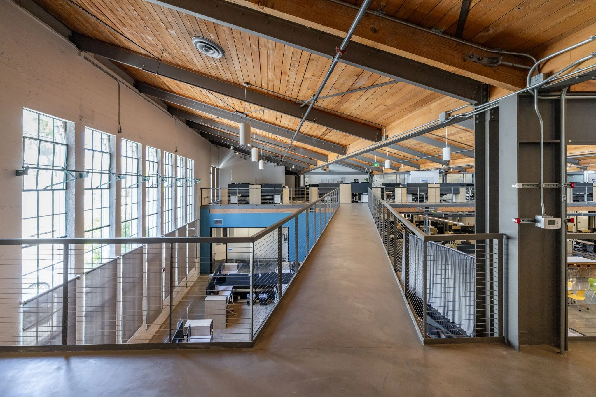 Industrial-style modern office space with a loft-style second floor, wooden ceiling, large windows, and open workstations.