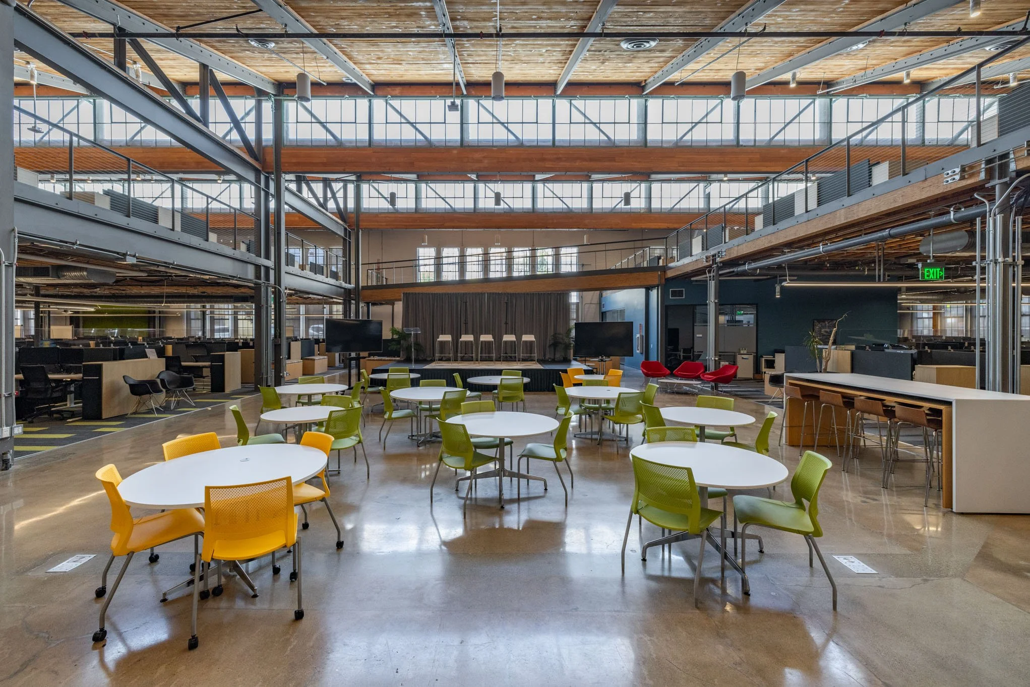 Open-plan modern office with round tables and colorful chairs, a stage with stools, and a multi-level interior with large windows and wooden accents.