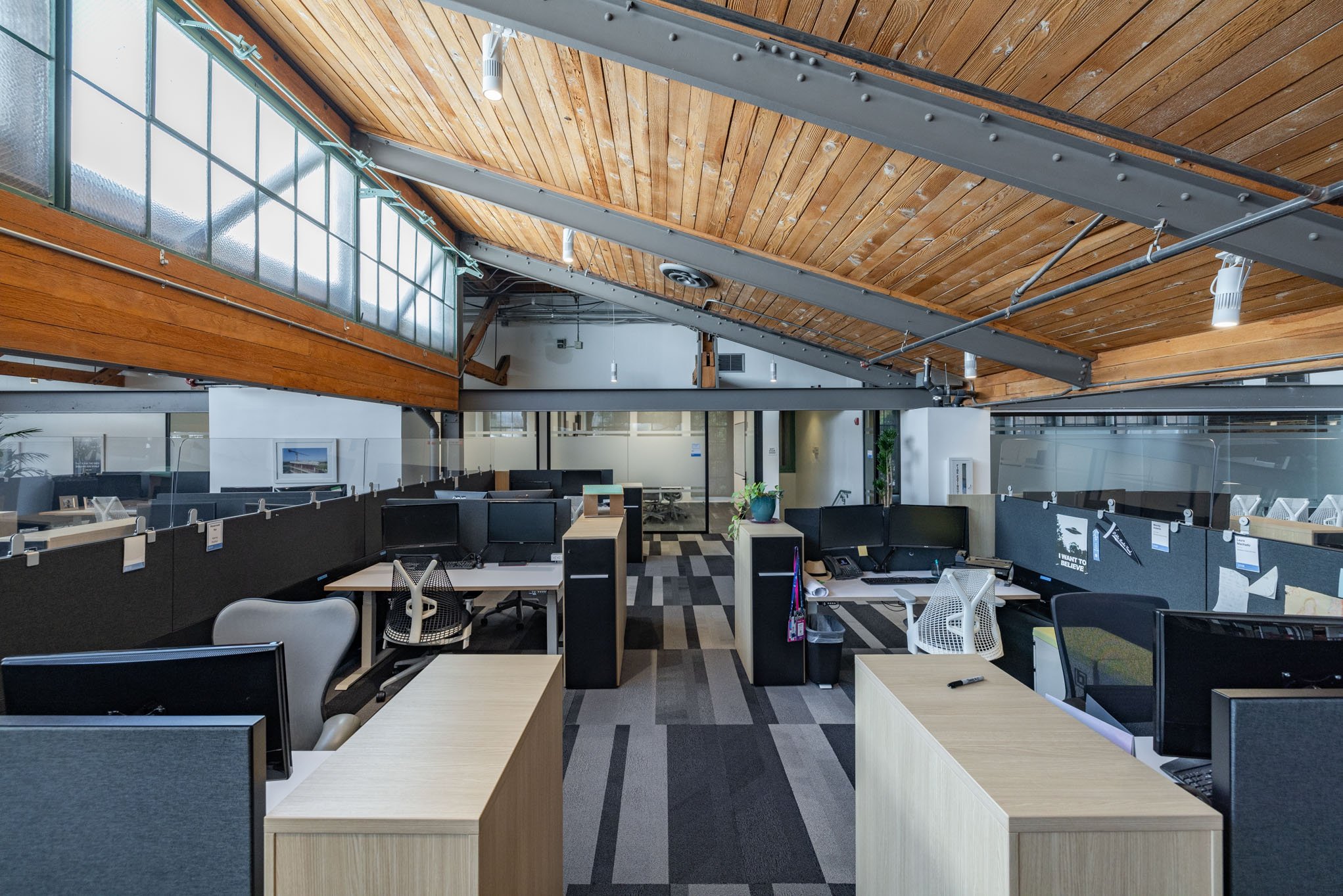 An empty modern office space with multiple desks and computers, glass partition walls, and a wooden ceiling with exposed beams.