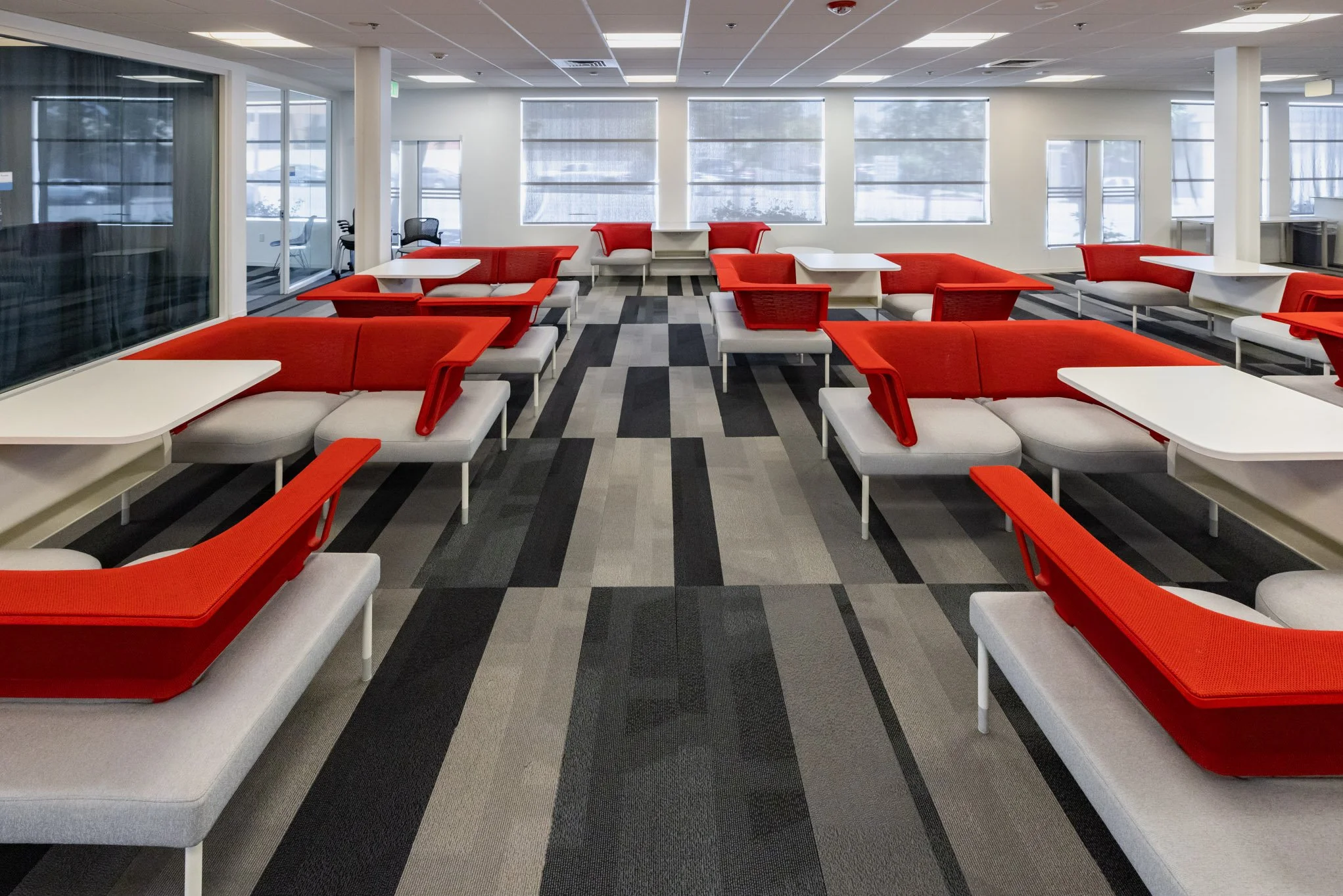 Empty lounge area with red and gray couches, white tables, gray and black patterned carpet, large windows, and glass doors in a modern building.