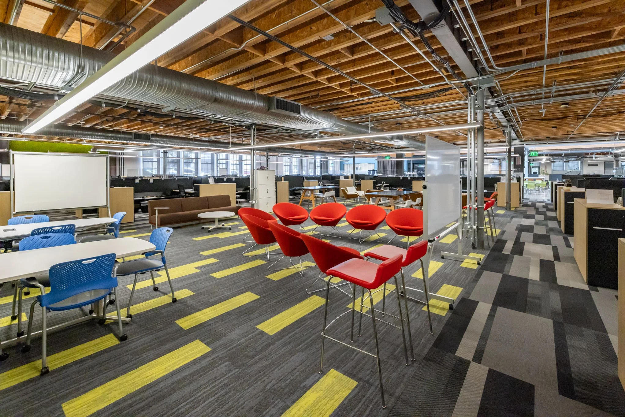 Modern open-plan office with colorful chairs, desks, and a whiteboard, featuring exposed wooden beams and ductwork on the ceiling.