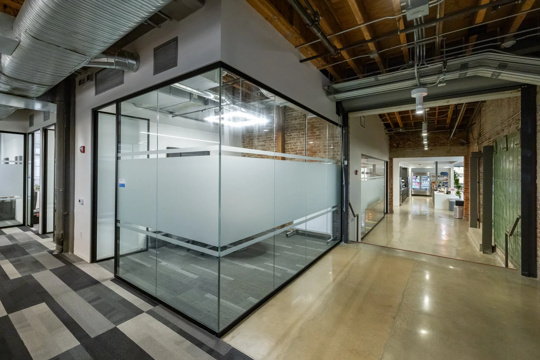 Modern office interior with glass-walled conference room and open workspace, exposed ductwork and brick walls.