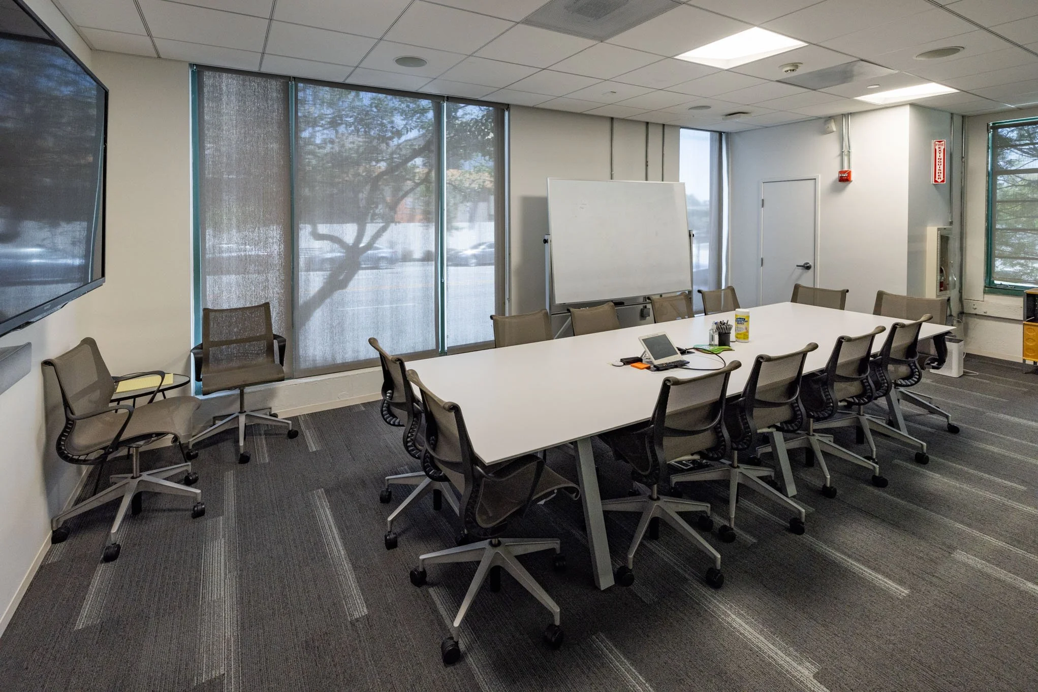 Empty conference room with a long white table, several office chairs, a whiteboard, a large television, and some office supplies on the table. Large windows with blinds are in the background.