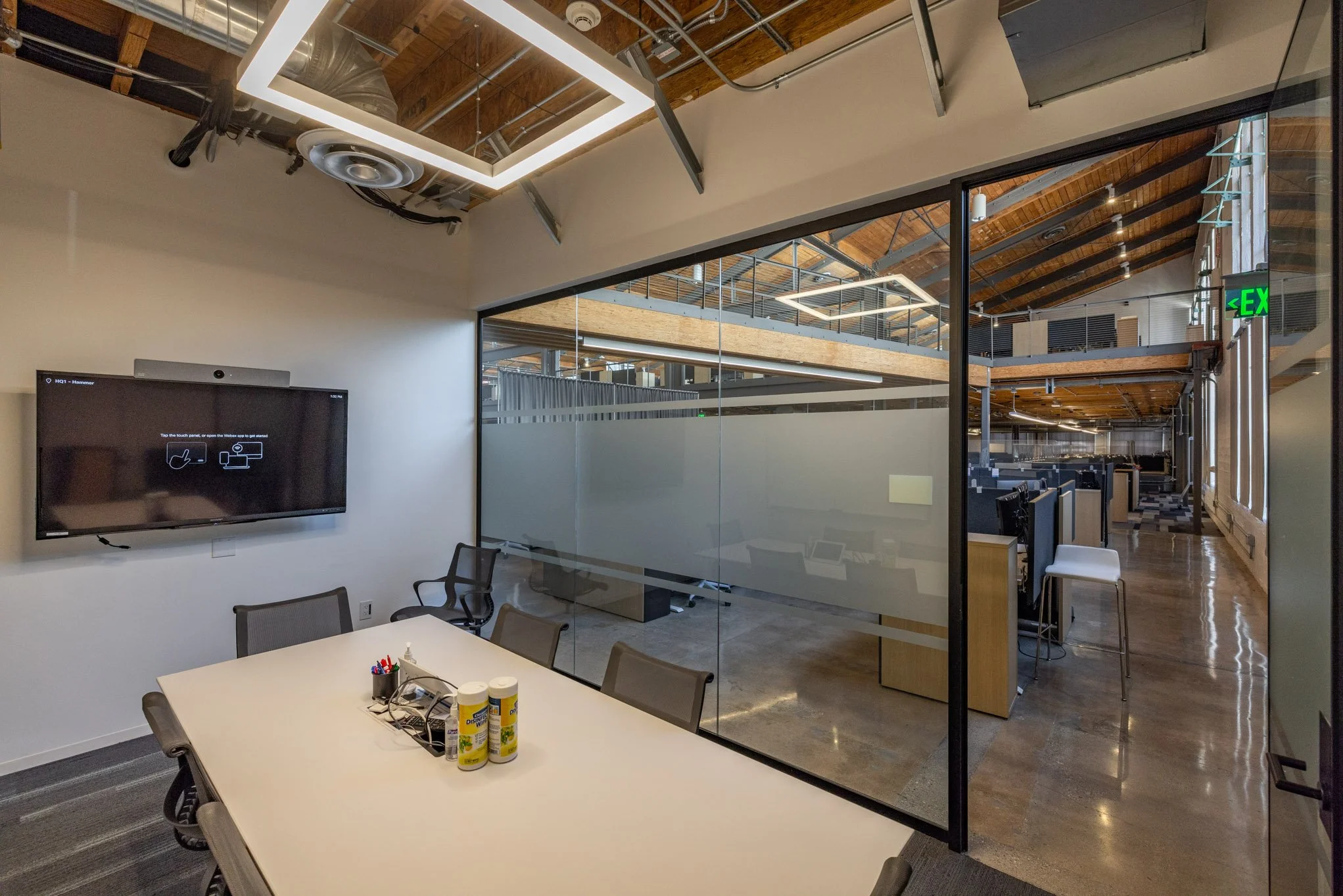 Modern office conference room with a white table, chairs, a wall-mounted screen displaying setup instructions, and a glass-walled office space beyond with cubicles and wooden accents.
