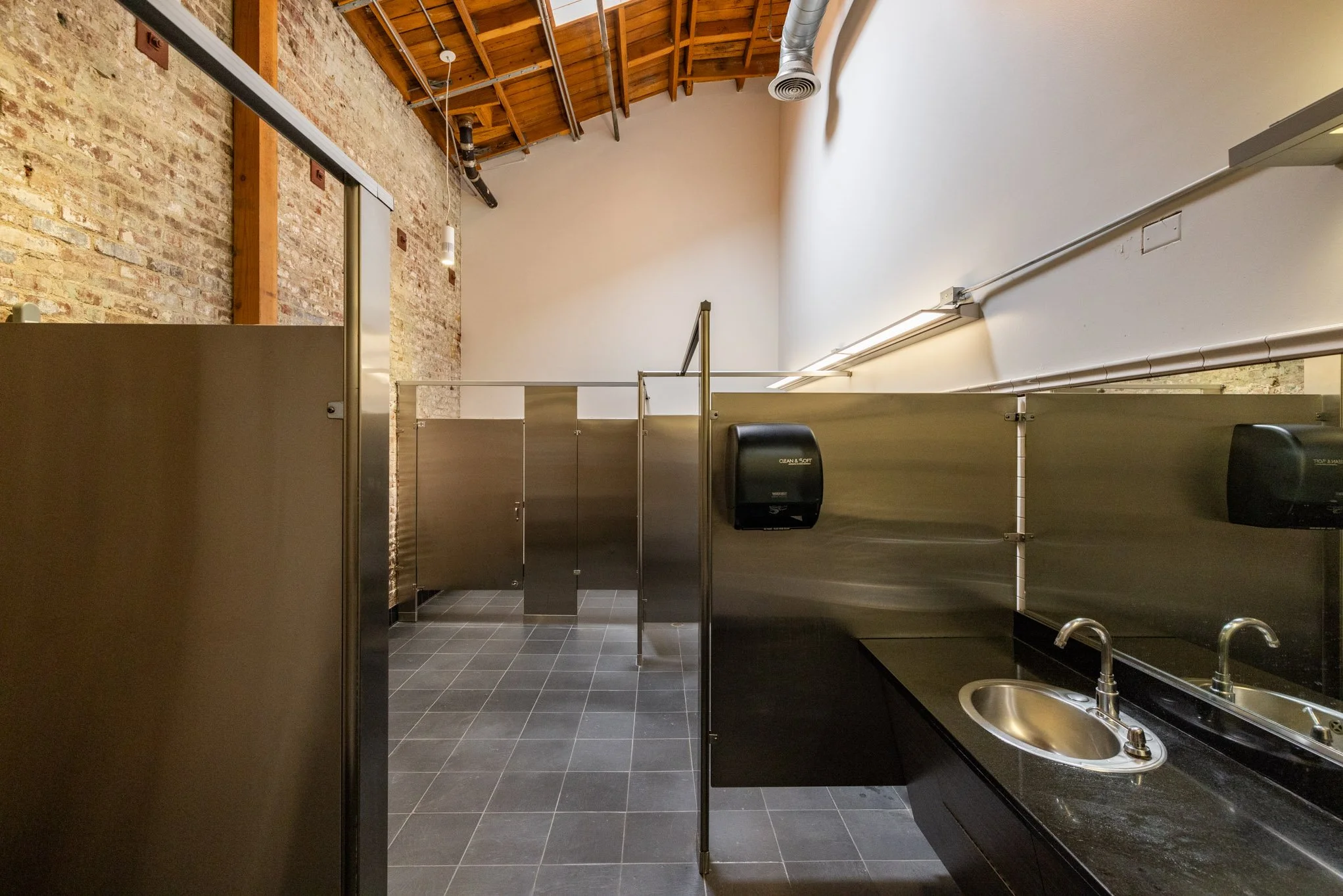 Public restroom with black countertops, stainless steel sink, soap dispensers, partitioned stalls, brick wall, and exposed wooden ceiling.