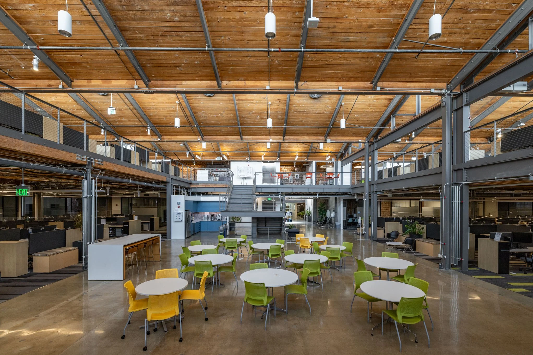 Modern open-plan office with a high wooden ceiling, metal beams, and bright lighting. Central area has round tables with green and yellow chairs, with workstations along the sides and a staircase leading to an upper level.