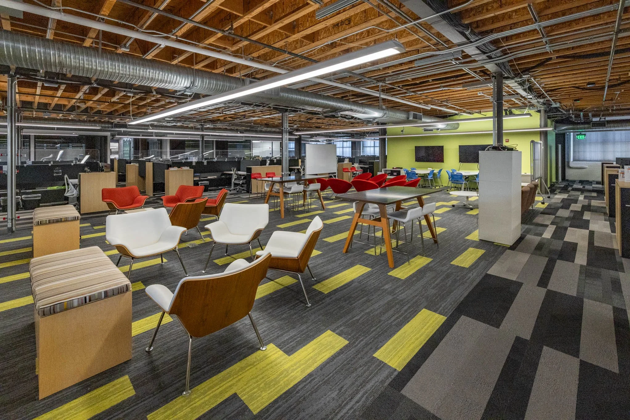 Modern office lounge area with various chairs, tables, and a green accent wall with two televisions, exposed ceiling with ductwork, and multiple workstations in background.