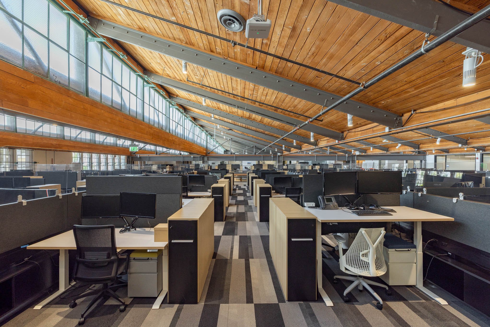 Modern office space with rows of workstations, computers, office chairs, and partitioned cubicles in a large open-plan room with a wooden ceiling, exposed beams, and glass windows.