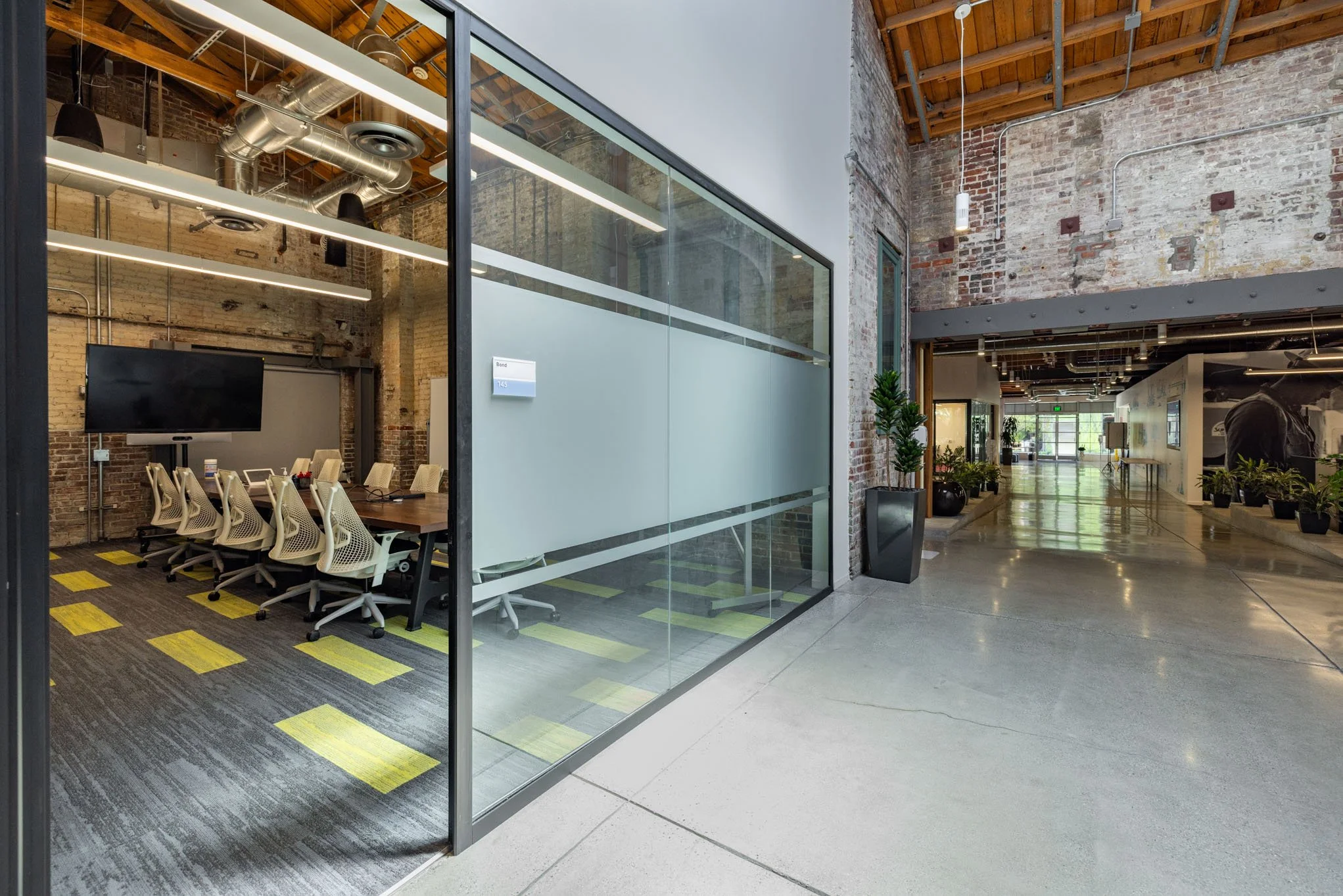 Modern office conference room with a large TV and beige chairs around a table, visible through a glass partition wall, with exposed brick walls and industrial style ceiling.
