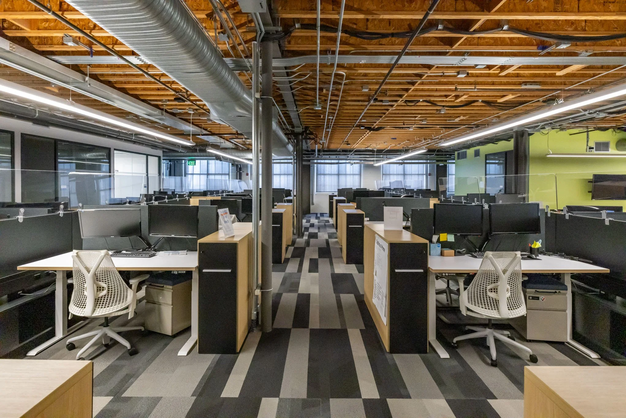 Empty office workspace with rows of desks, chairs, and computer monitors, separated by black partitions, under a ceiling with exposed wooden beams and industrial ducts.