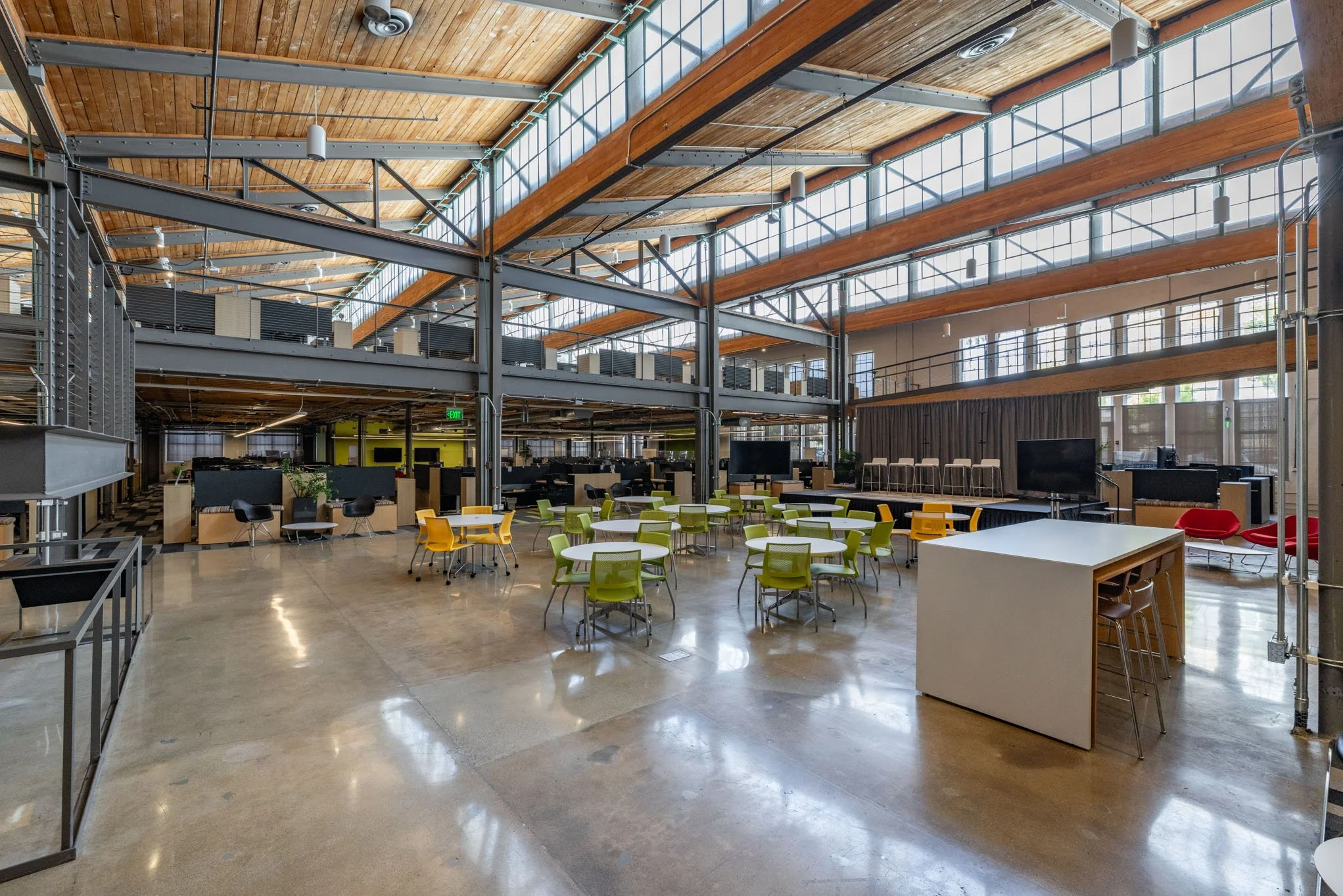 A spacious, modern co-working space with high wooden ceilings, large windows, green and yellow chairs, white tables, a few red chairs, and multiple large screens.