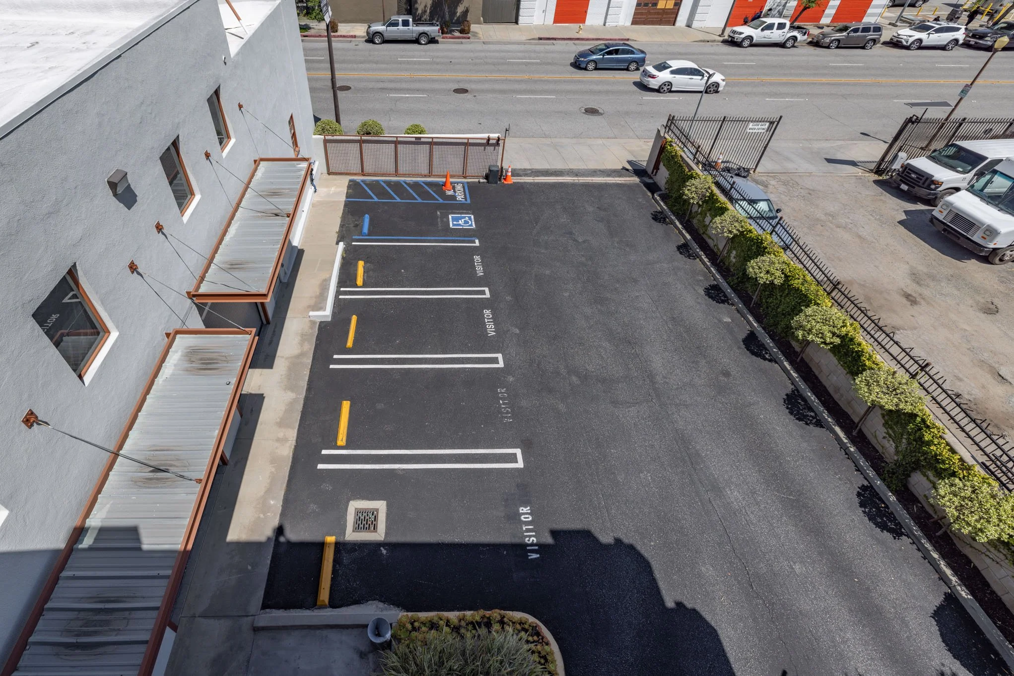 Overhead view of a parking lot with designated spaces, including handicap accessible and visitor spots, next to a gray building with awnings. Cars are parked across the street, and there is a row of small bushes along the right side