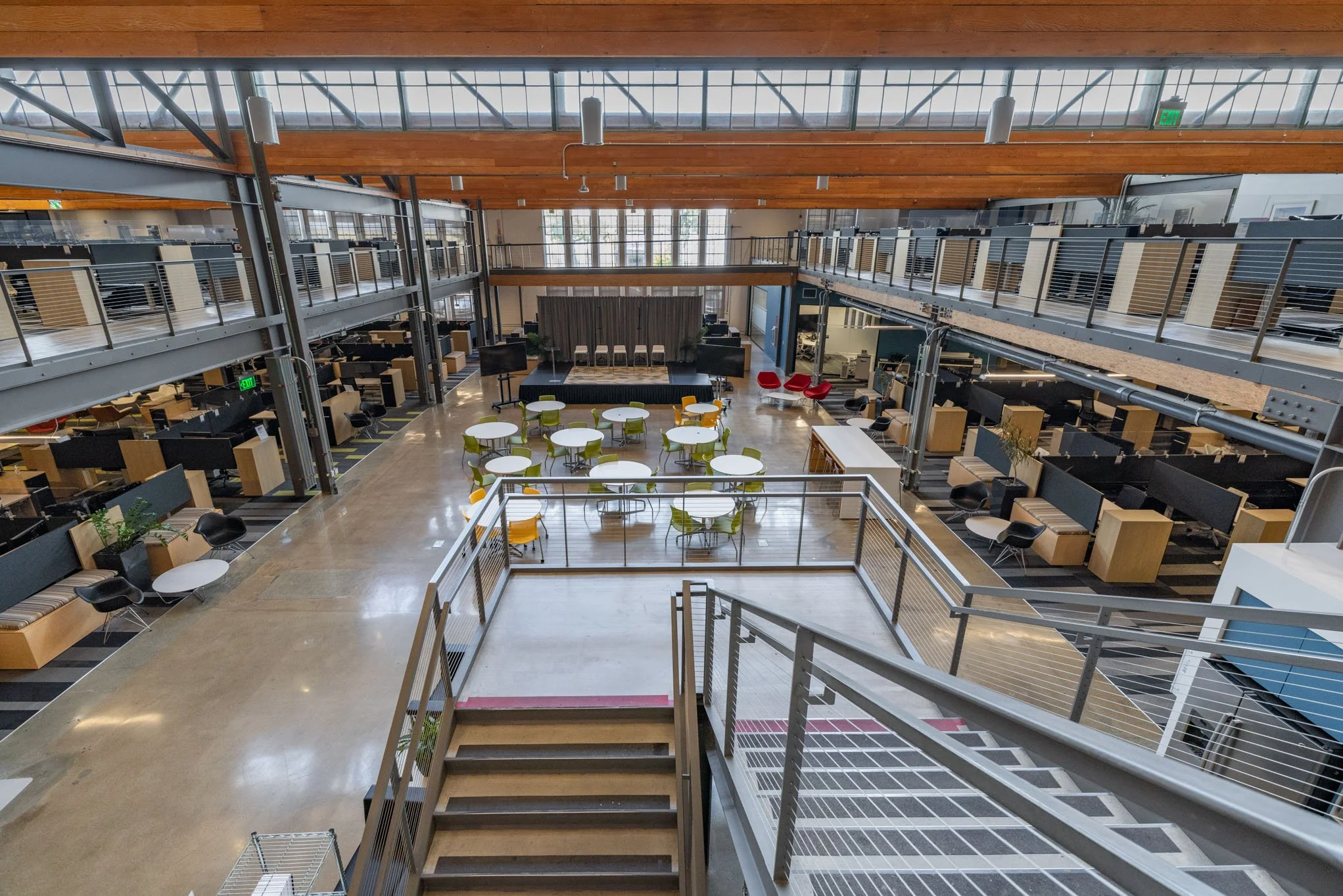 An open and modern office space with a mezzanine level, featuring tables with green and yellow chairs, booths along the walls, and a stage at the back with curtains and twin pianos, illuminated by large windows and ceiling lights.