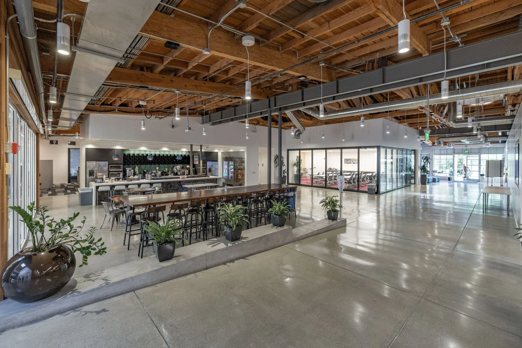 Modern indoor space with high wood ceiling, polished concrete floor, a coffee station, a glass-enclosed conference room, and seating areas with plants.