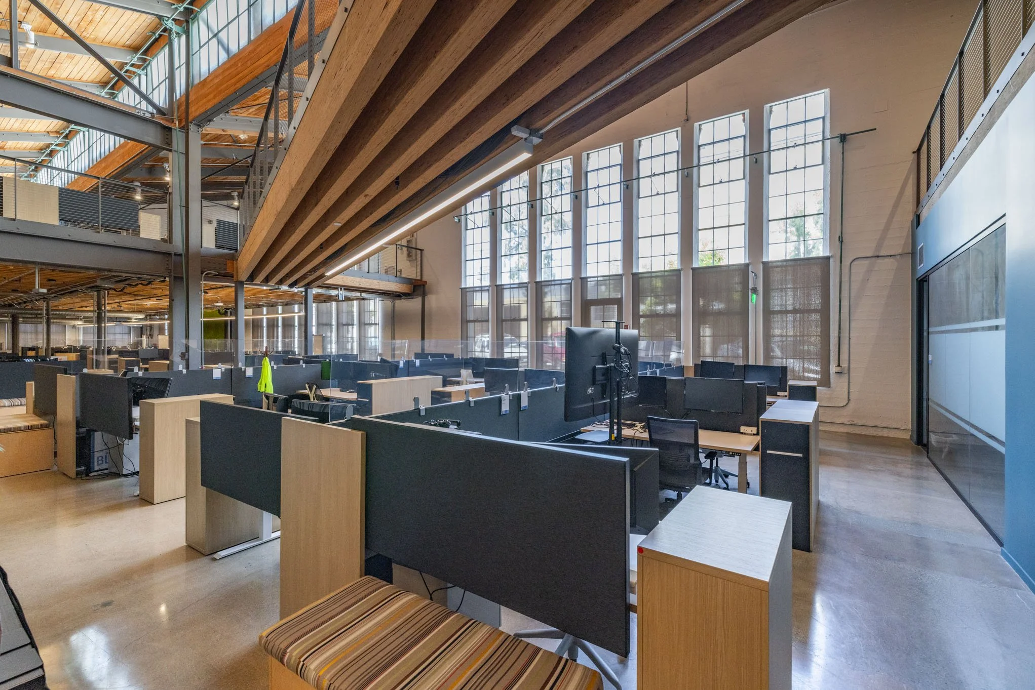 Open office space with workstations, cubicles, and large windows, featuring high ceilings with wooden beams