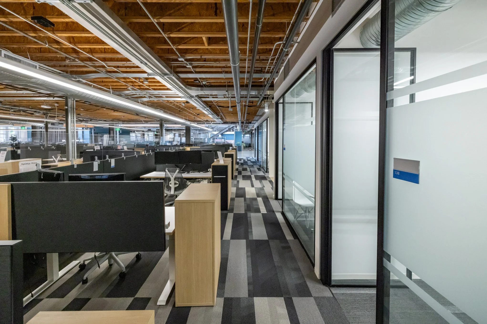 Modern office with workstations, glass-walled private offices, and exposed wooden ceiling in an open-plan layout.