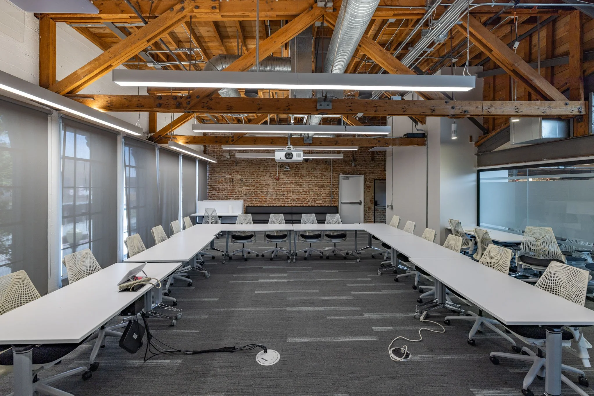 Empty modern conference room with U-shaped white tables, white ergonomic chairs, exposed wooden beams, brick wall, large windows with blinds, ceiling projectors, and electronic equipment on the floor.