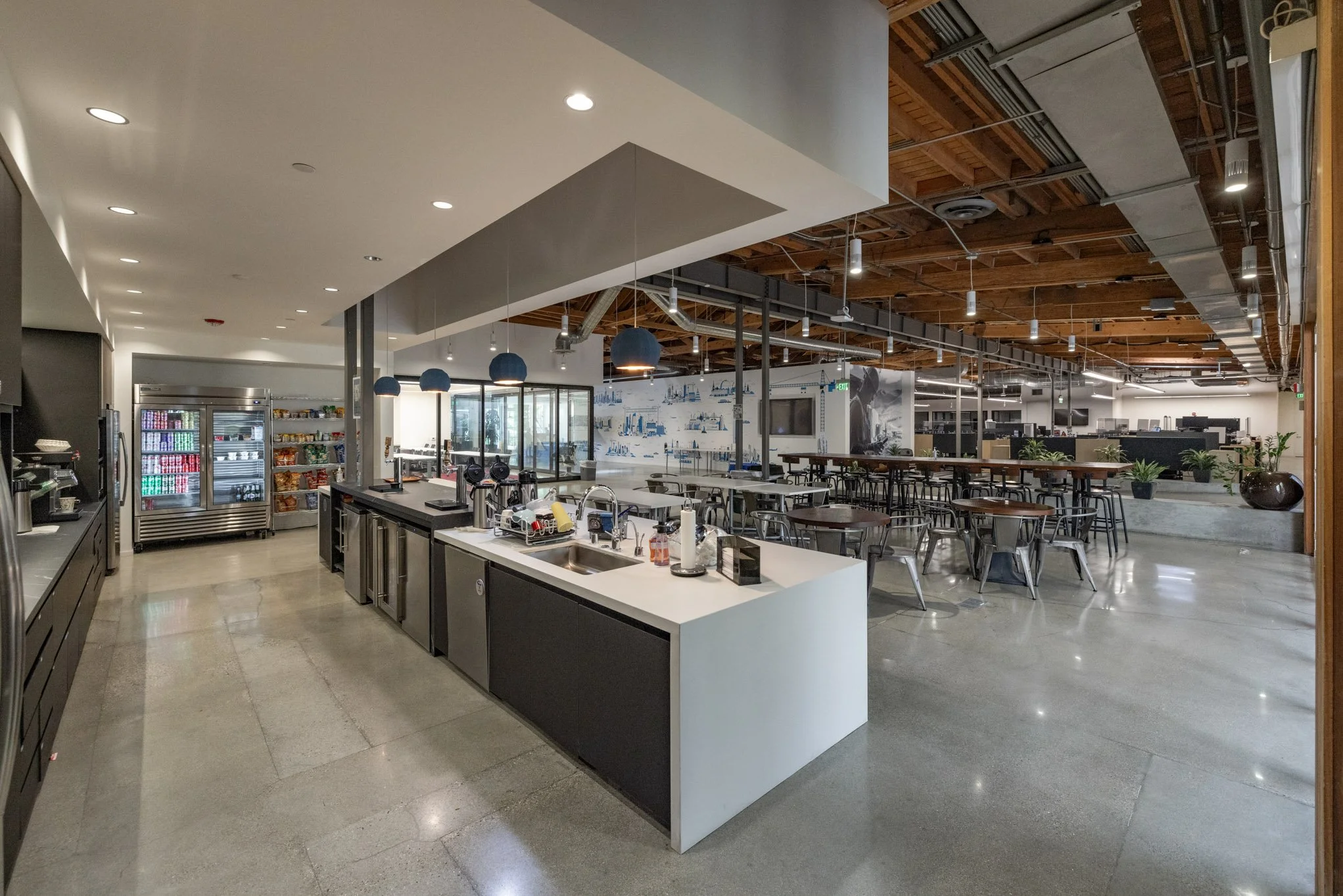 Modern café or communal dining space with a kitchenette area, tables, chairs, and potted plants in a spacious, industrial-style environment.