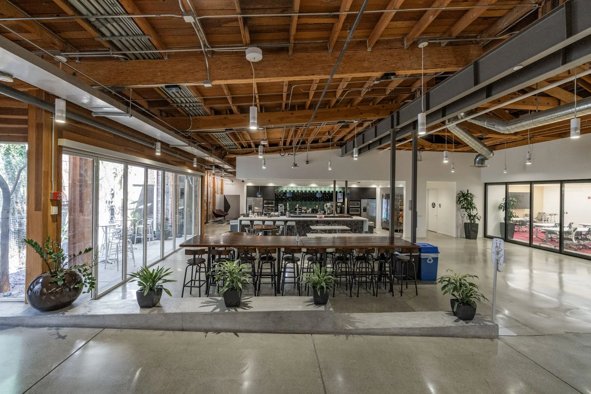 Interior of a modern cafe or dining area with large windows, wooden ceiling, and a central bar area. There are potted plants near the entrance and seating arrangements around the space, including a long wooden table with stools and additional seats b