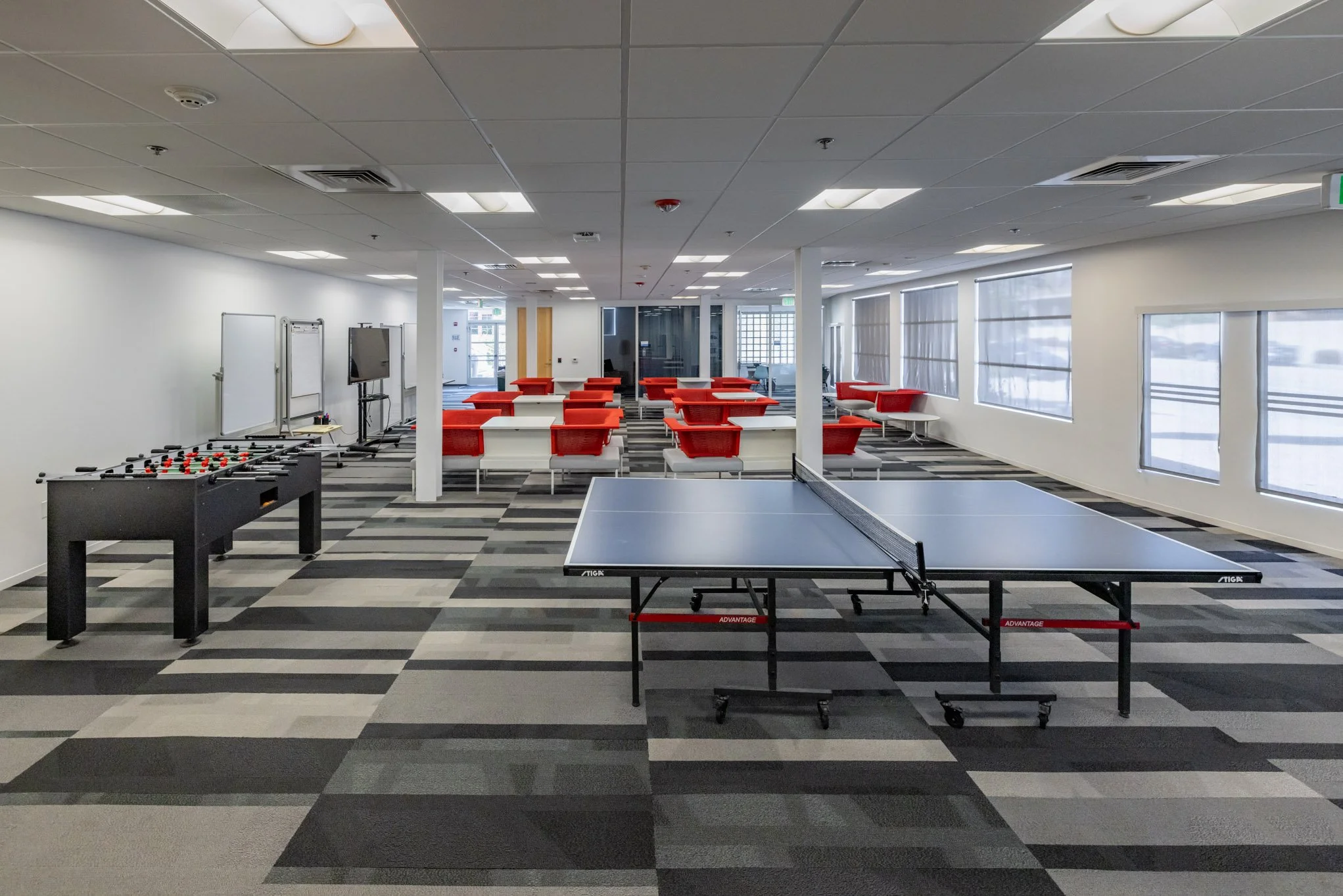 An empty game room with a foosball table, a ping pong table, a television, whiteboards, and red and white seating area, all illuminated by large windows and overhead lighting.