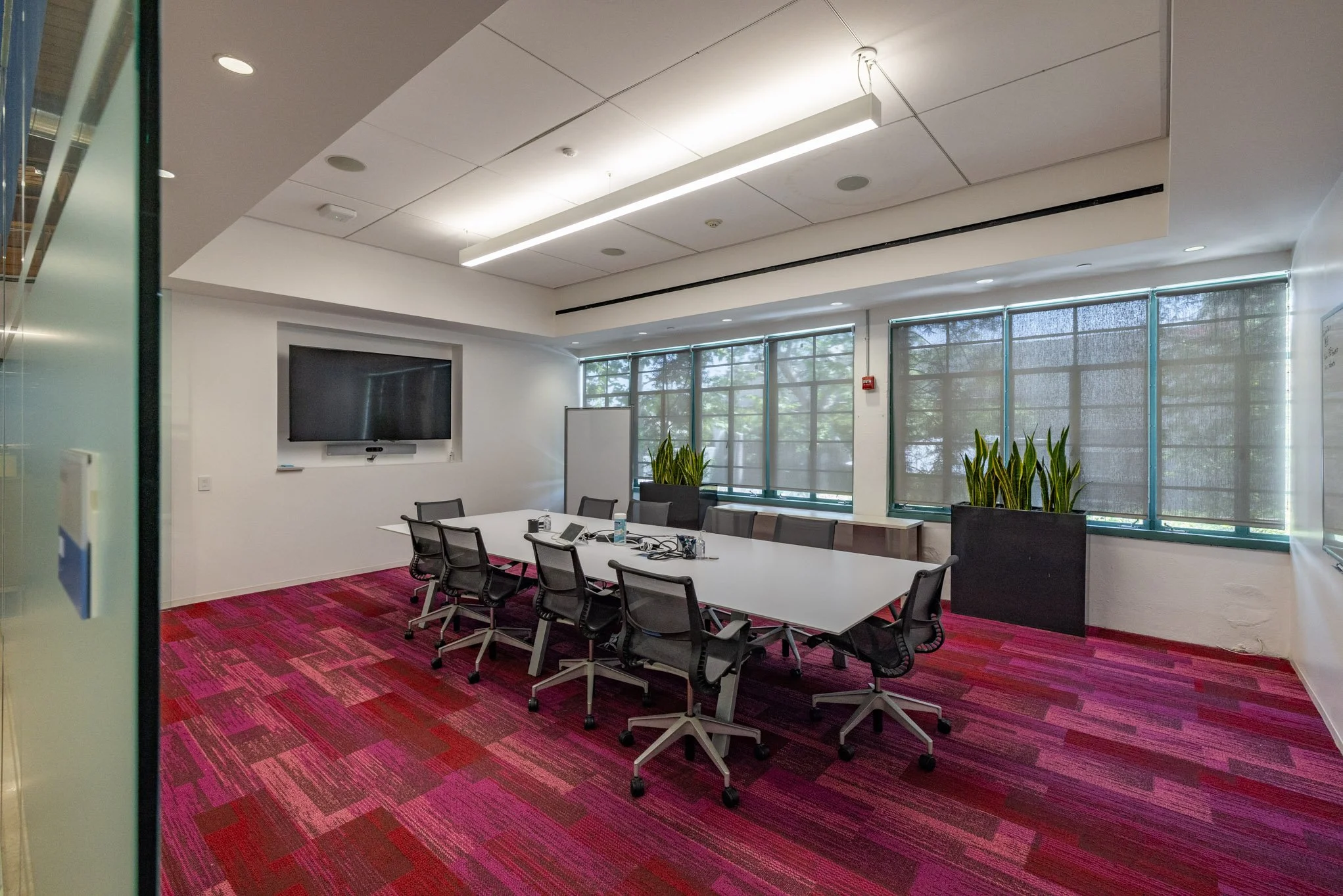 Empty conference room with a white table, black chairs, large windows with shades, plants, and a wall-mounted television.