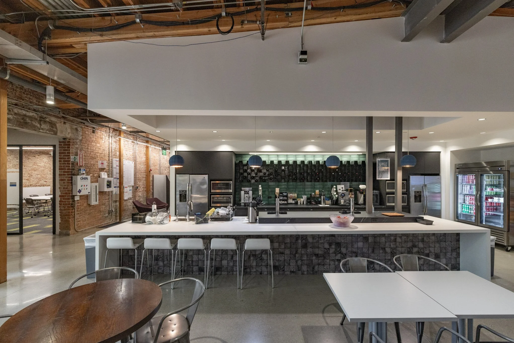 Modern cafe counter with bar stools, coffee machines, and refrigerated beverage display in a contemporary industrial-style space with exposed brick walls and wood ceiling beams.