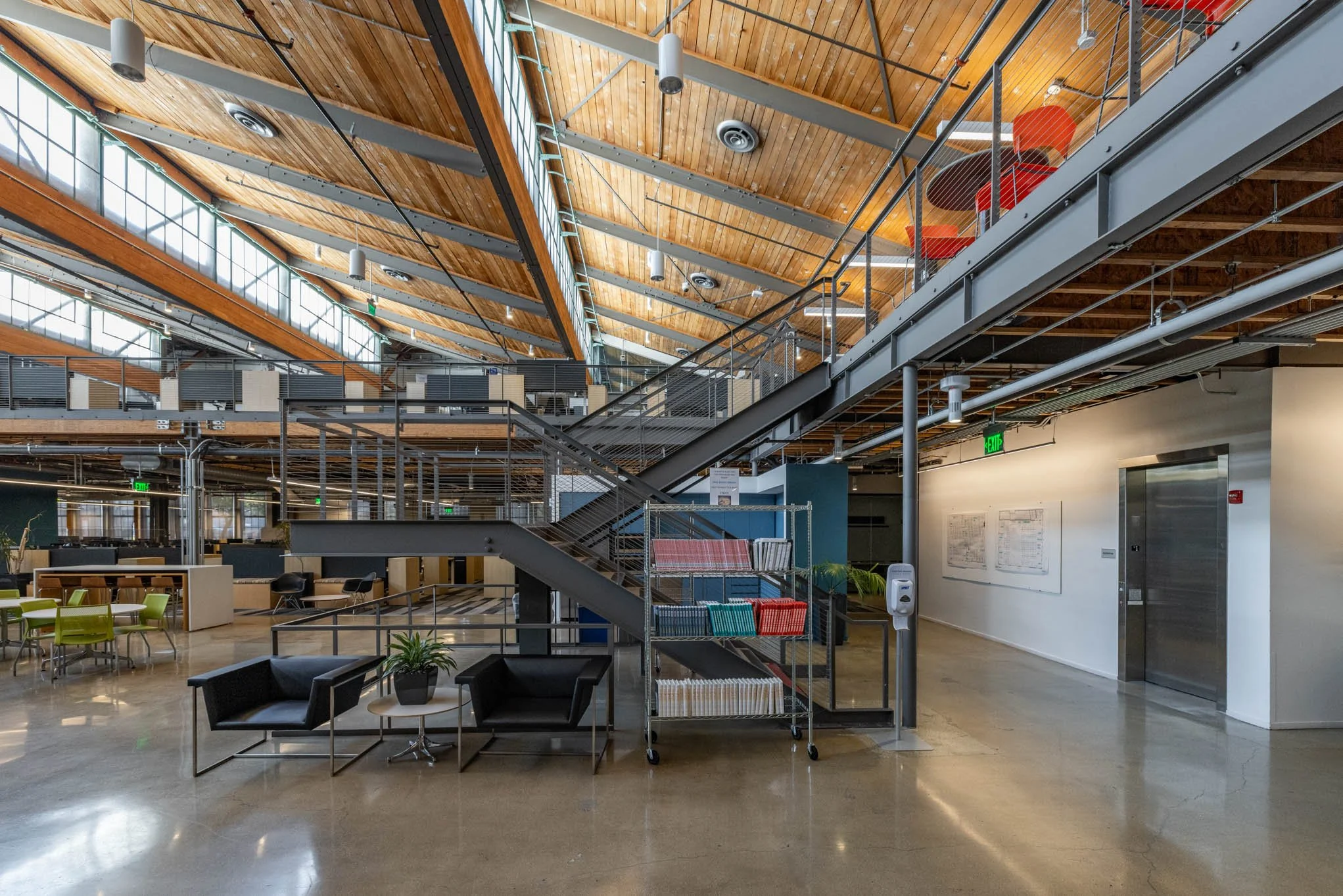 Interior view of a modern office with a high wooden ceiling, large windows, metal staircase, and seating area with chairs, couches, and plants.