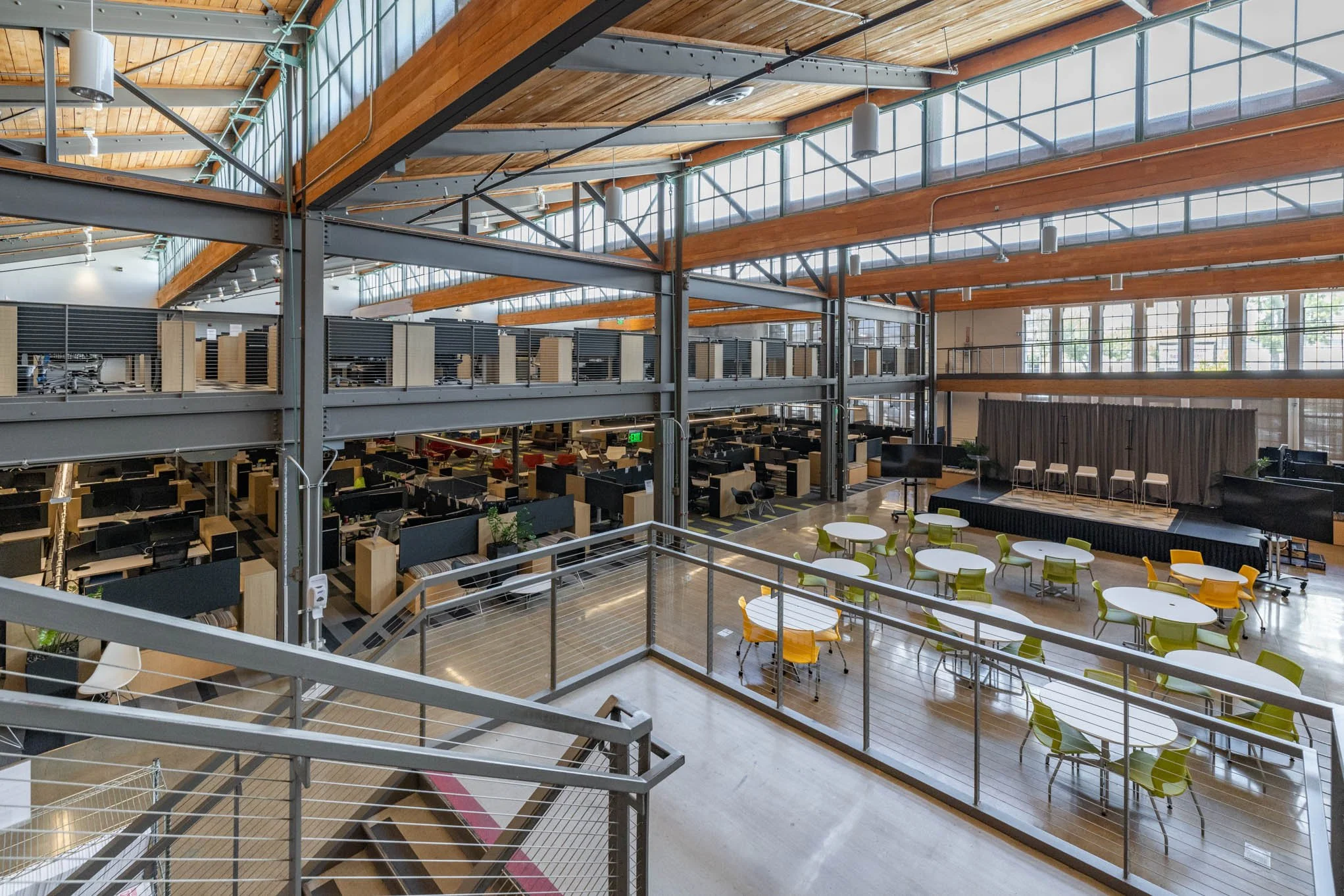 Interior of a modern, spacious office building with an open floor plan, multiple workspaces with cubicles, an elevated lounge area, and a stage with chairs and tables. The ceiling features exposed beams and large windows allow natural light to fill t