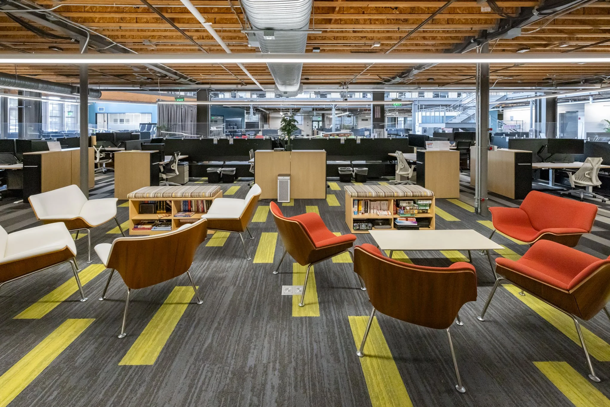 Modern open-plan office space with chairs and small tables in the foreground, cubicles and desks in the background, exposed ceiling with wood and metal beams, and large windows allowing natural light.