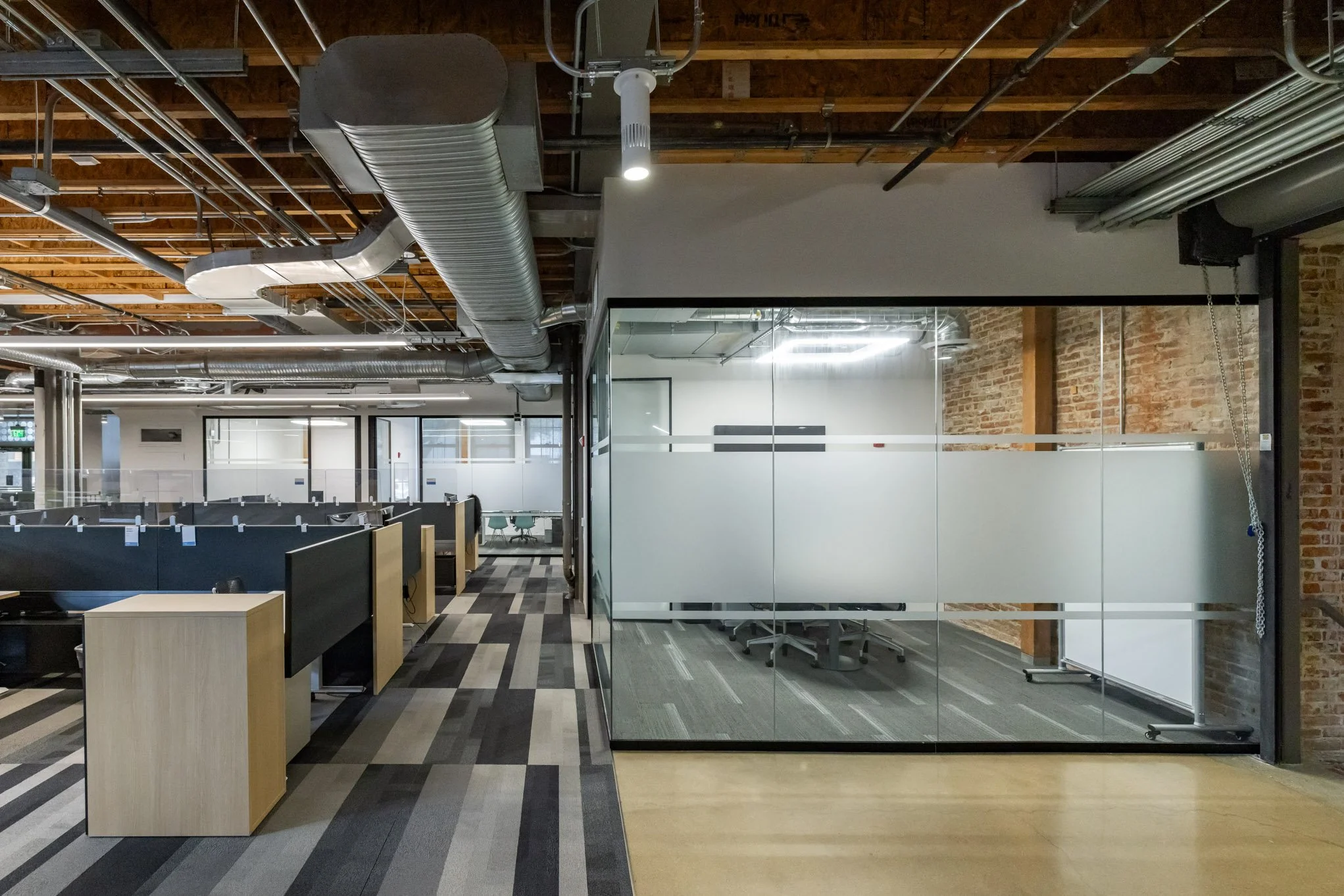 Modern office space with cubicles on the left and a glass-walled conference room on the right. Exposed ductwork and wooden ceiling beams create an industrial look.