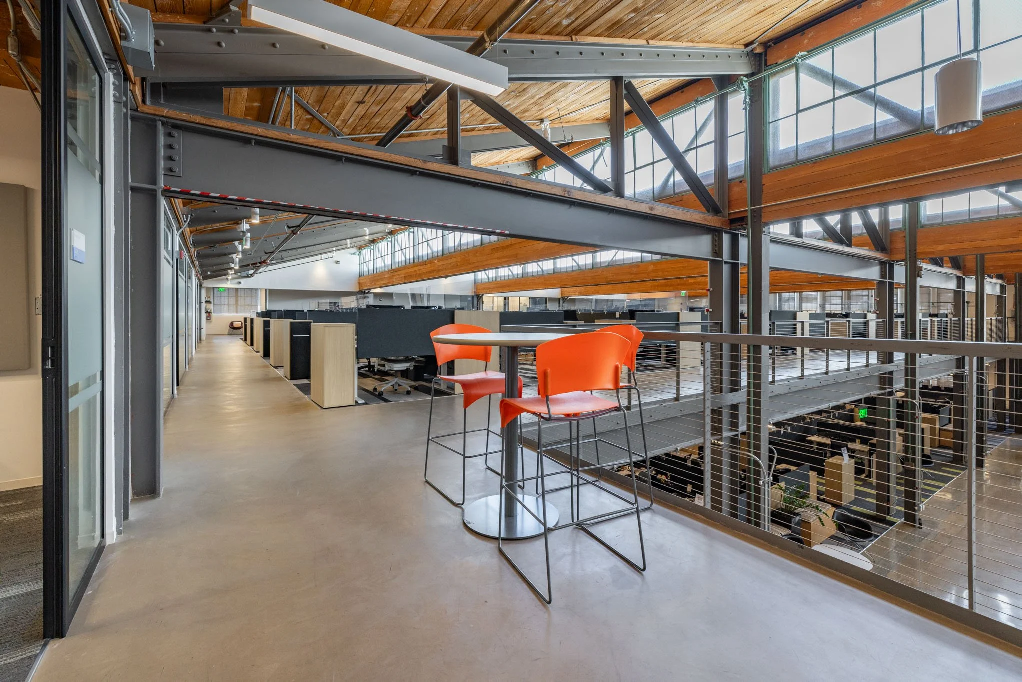 Modern office balcony with orange chairs and a small round table, overlooking a two-story open workspace with cubicles and glass windows, featuring wooden ceiling beams and metal framing.