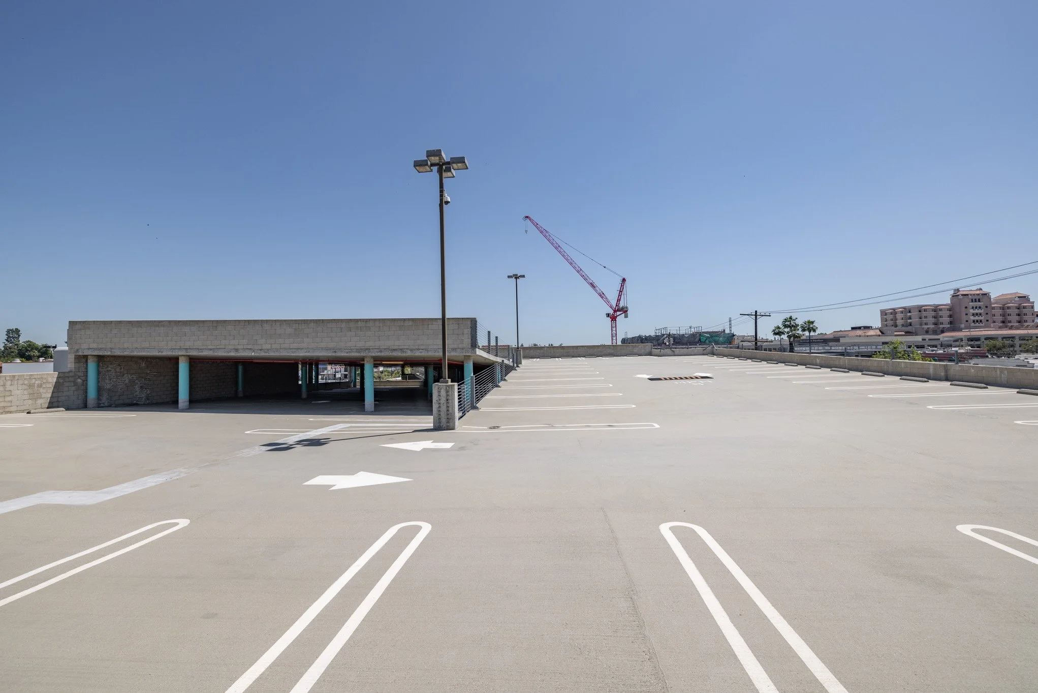 Empty rooftop parking lot with white painted parking space lines, a small parking garage structure on the left, a construction crane in the background, and some buildings and utility poles under a clear blue sky.