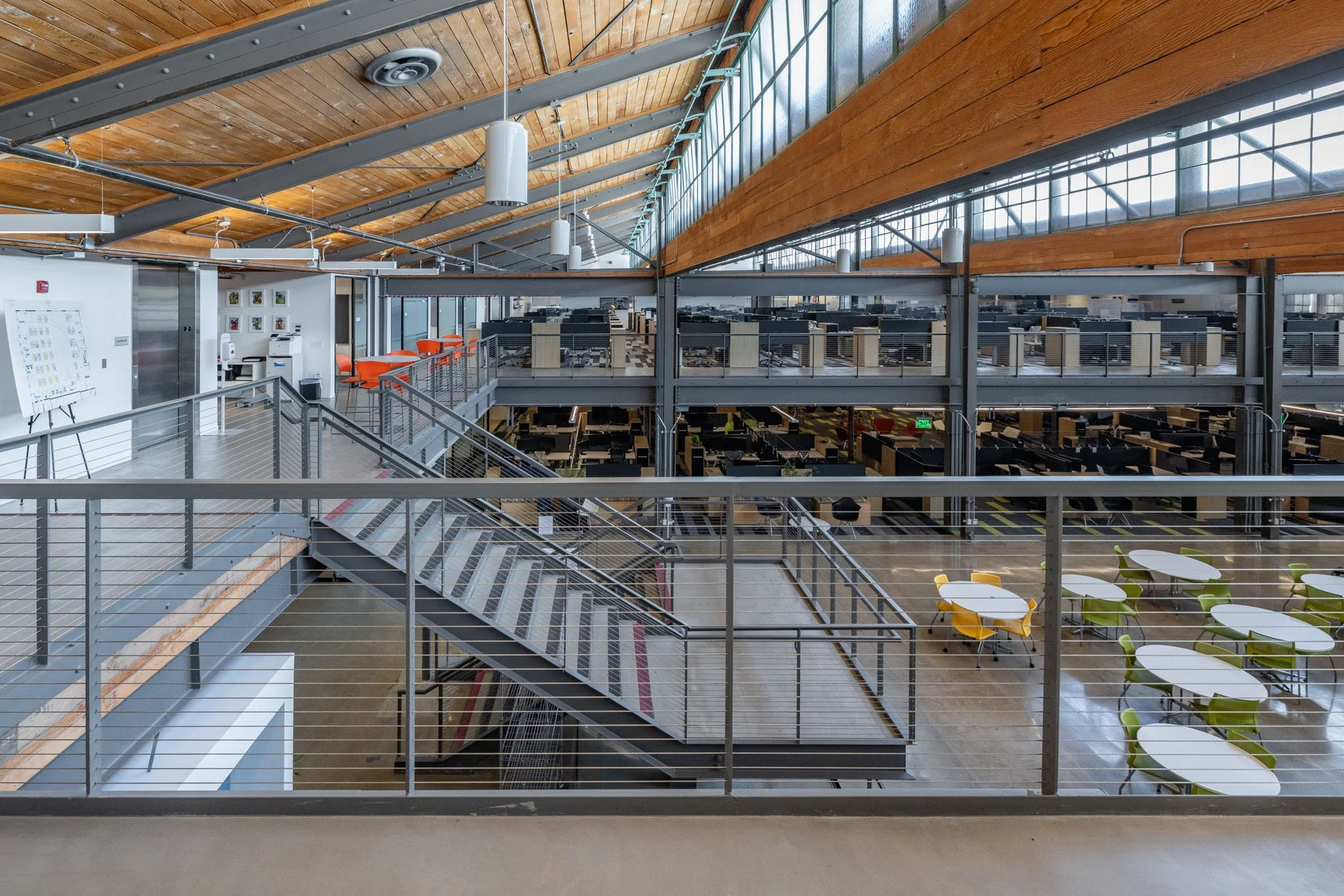 Interior view of a modern office with an open-plan layout, featuring multiple workstations on two levels, a staircase, round tables with chairs, and a high wooden ceiling with large windows.