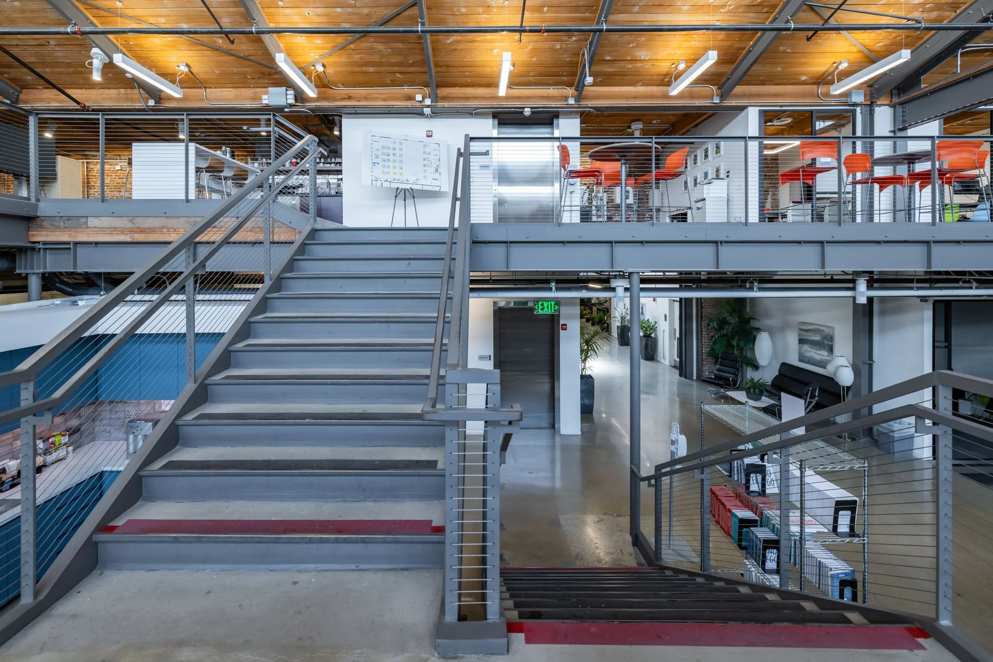 Interior view of a modern office with metal staircase, mezzanine level with orange chairs and desks, and a hallway with black seating and plants.